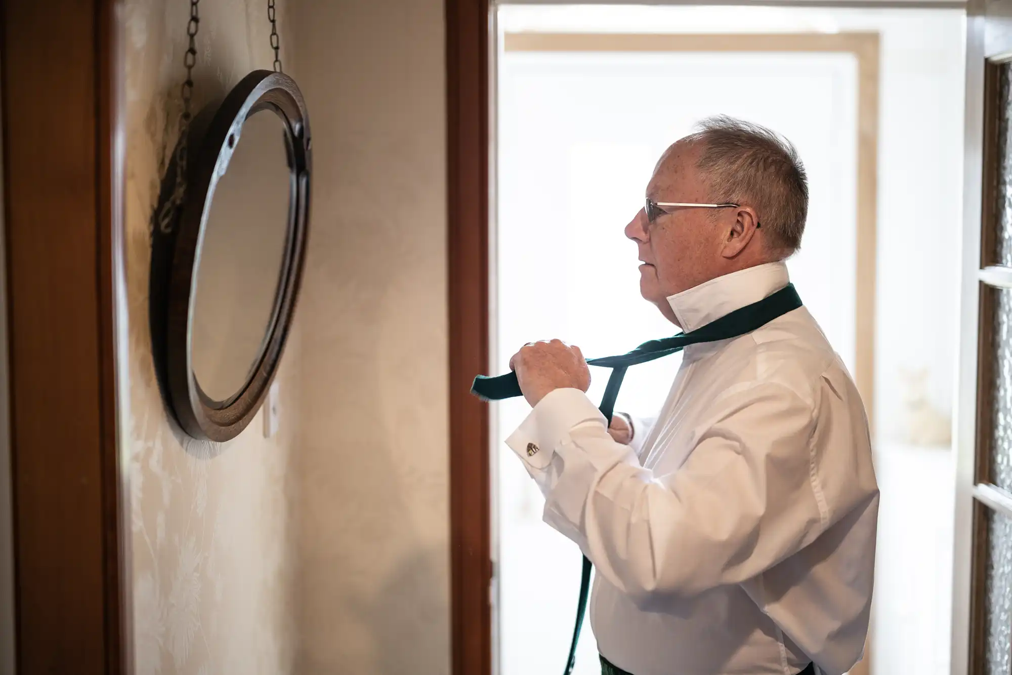 An elderly man wearing a white dress shirt adjusts his green tie while looking into a round wall mirror.