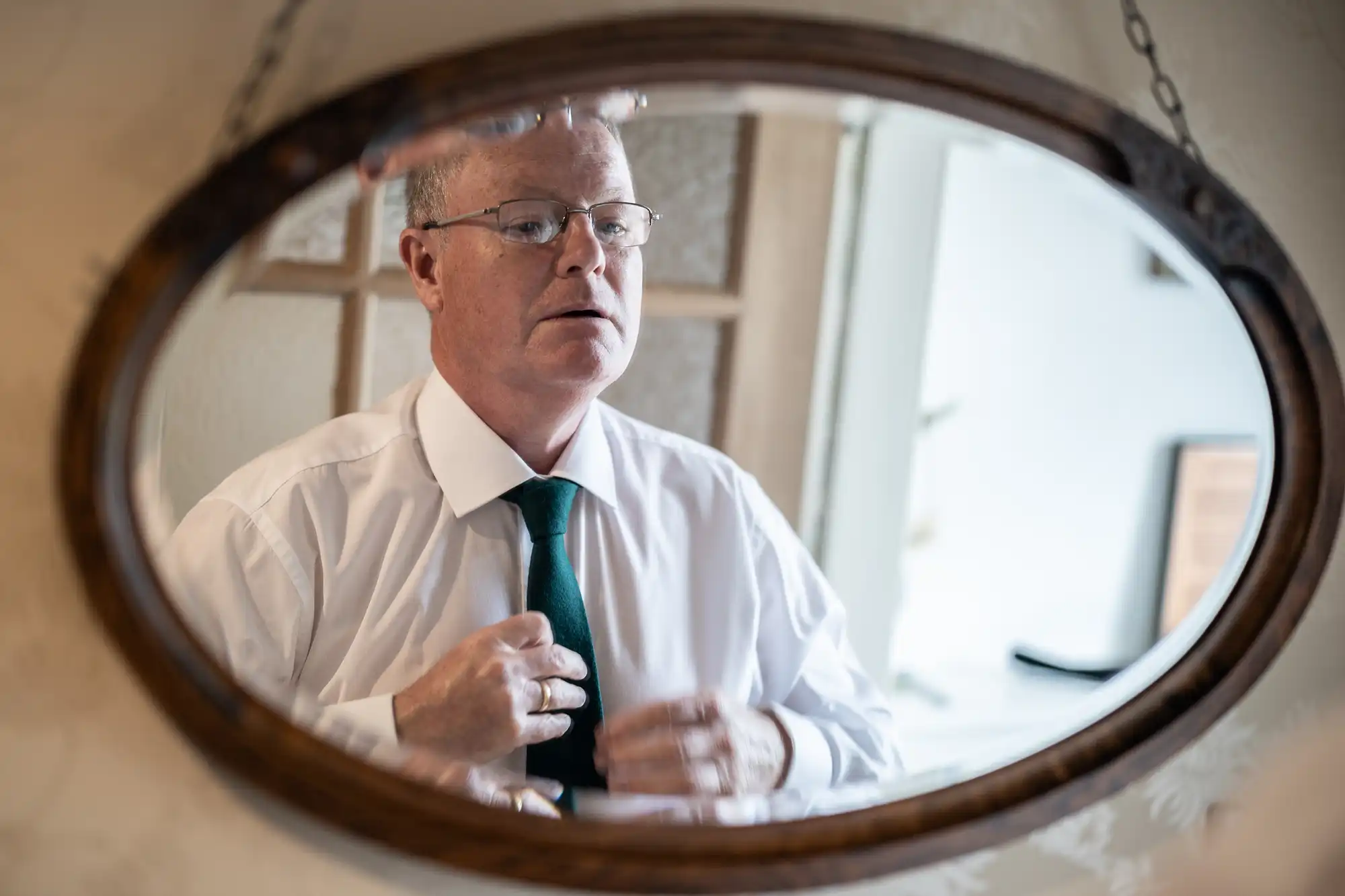 A man is adjusting his tie while looking into an oval mirror. He is wearing a white shirt and green tie.