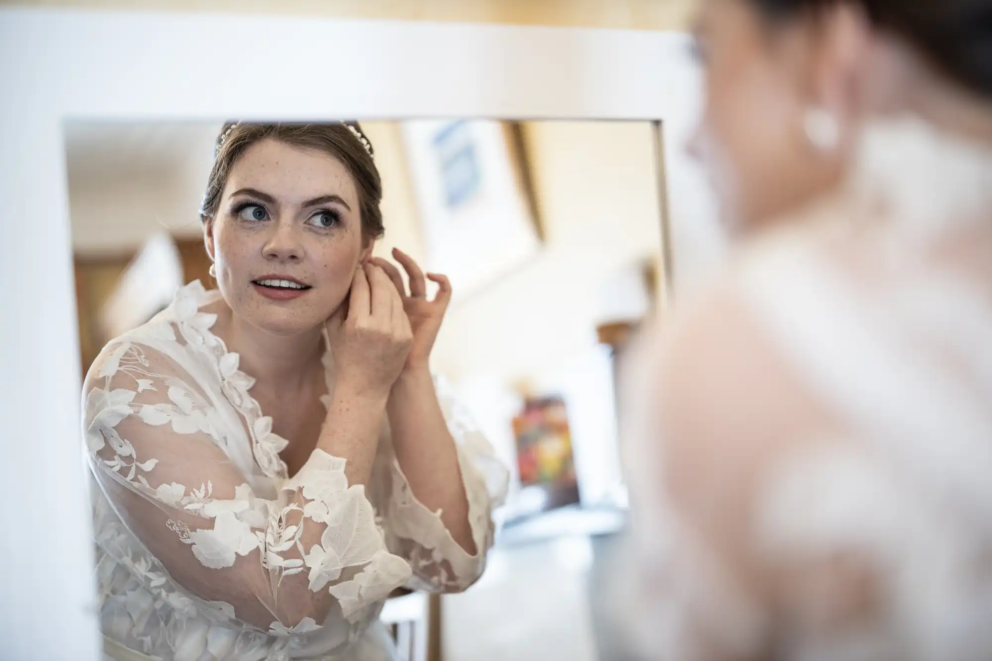 A bride in a white floral lace dress looks in a mirror while putting on earrings.