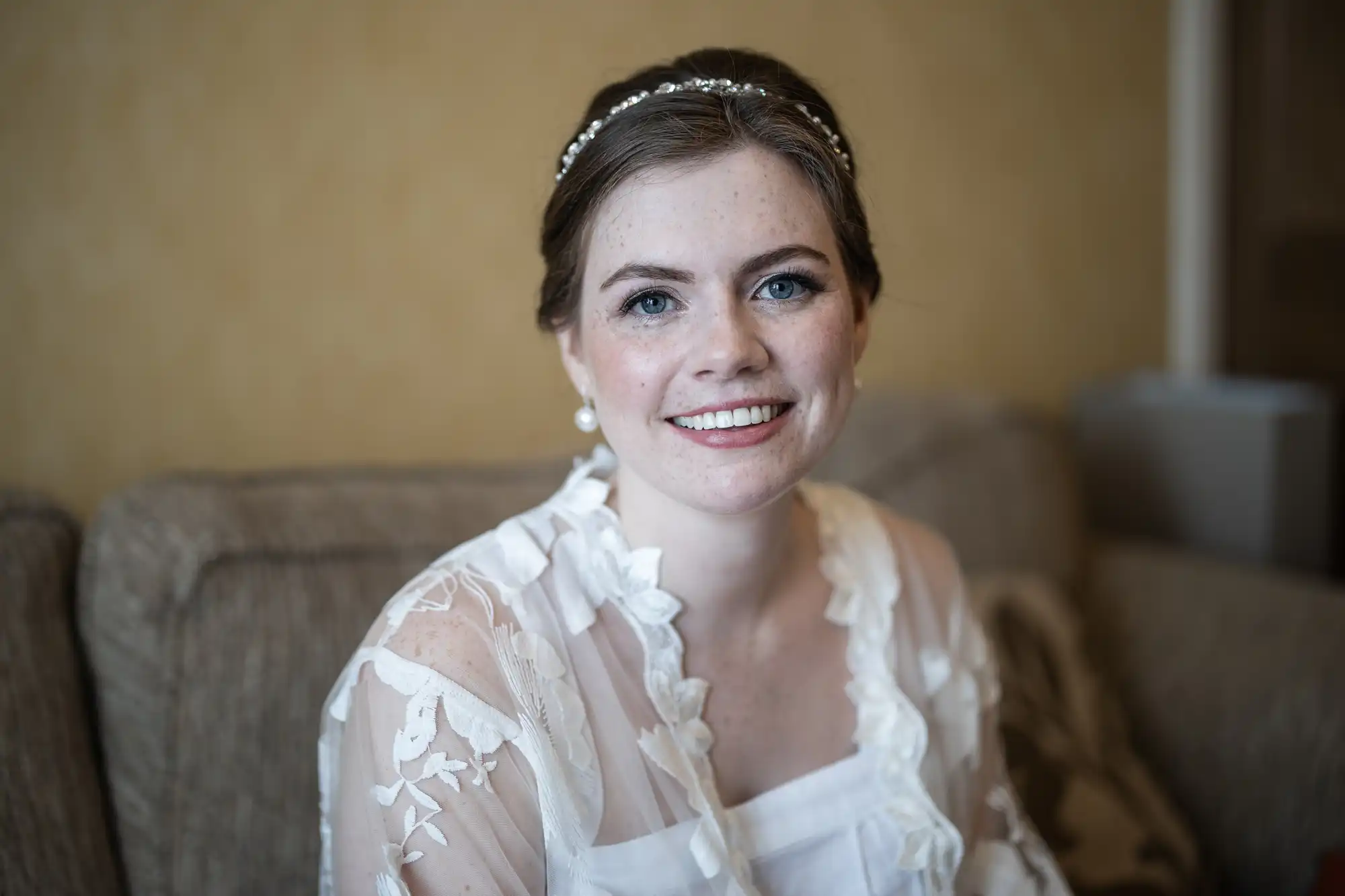 A woman with dark hair in a bun, wearing a white lace dress and pearl earrings, smiles while sitting on a beige couch with a light brown background.