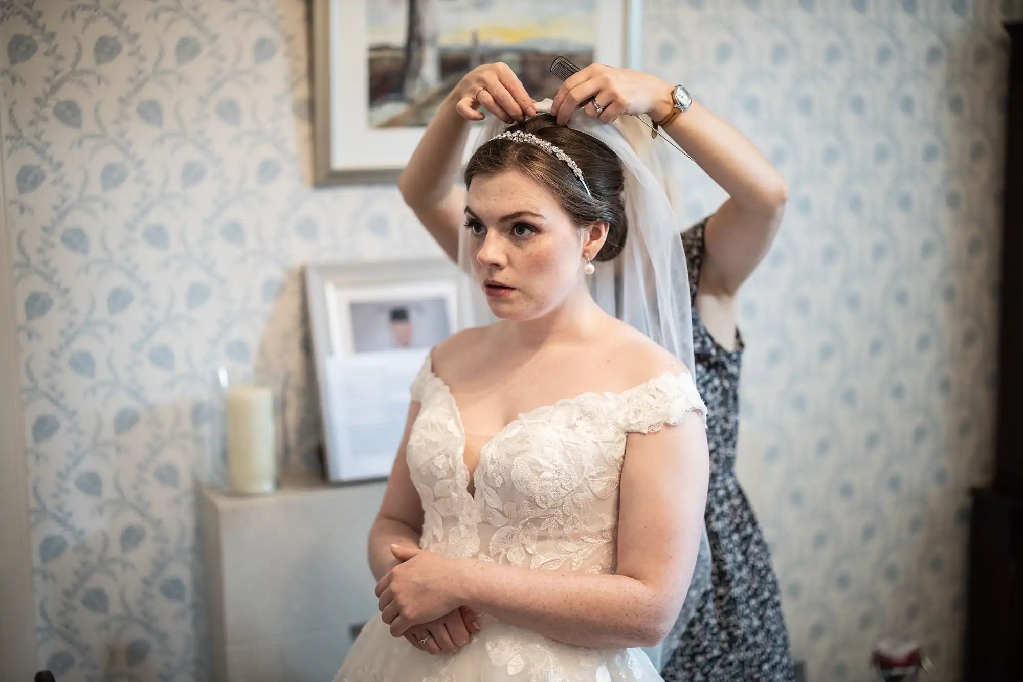 A bride wearing a white off-shoulder lace gown stands as another person adjusts her headpiece in a room with a framed picture on the wall and a candle on a table.