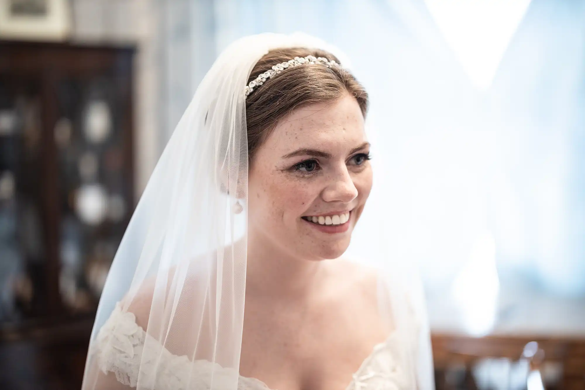 A bride, wearing a white dress and veil with a headband, smiles indoors with soft lighting and a blurred background.