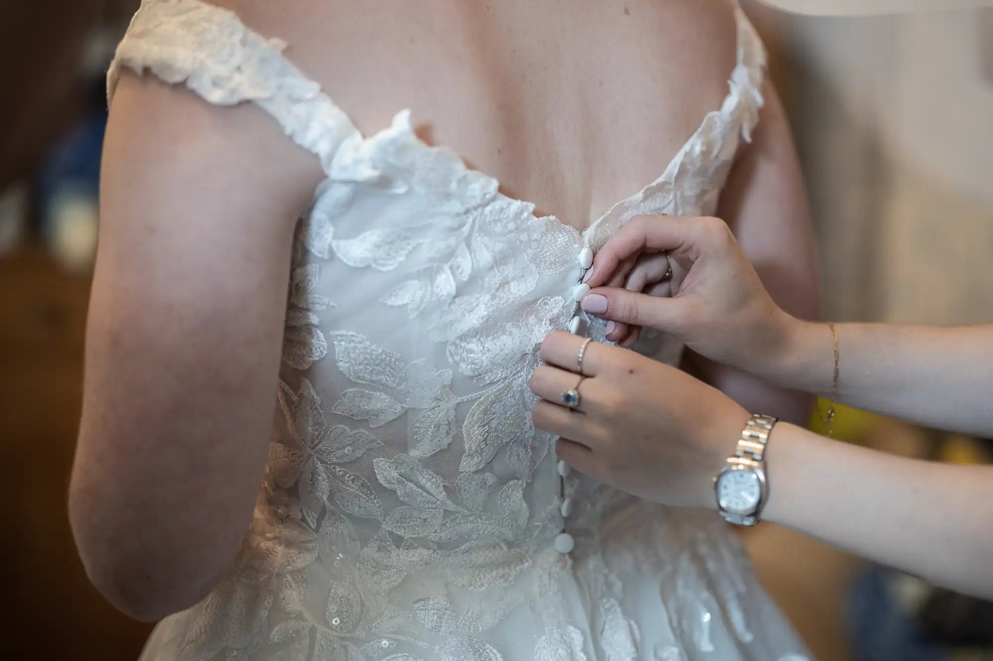 Close-up of hands fastening buttons on the back of a lace wedding dress worn by a person with a watch on their wrist assisting.