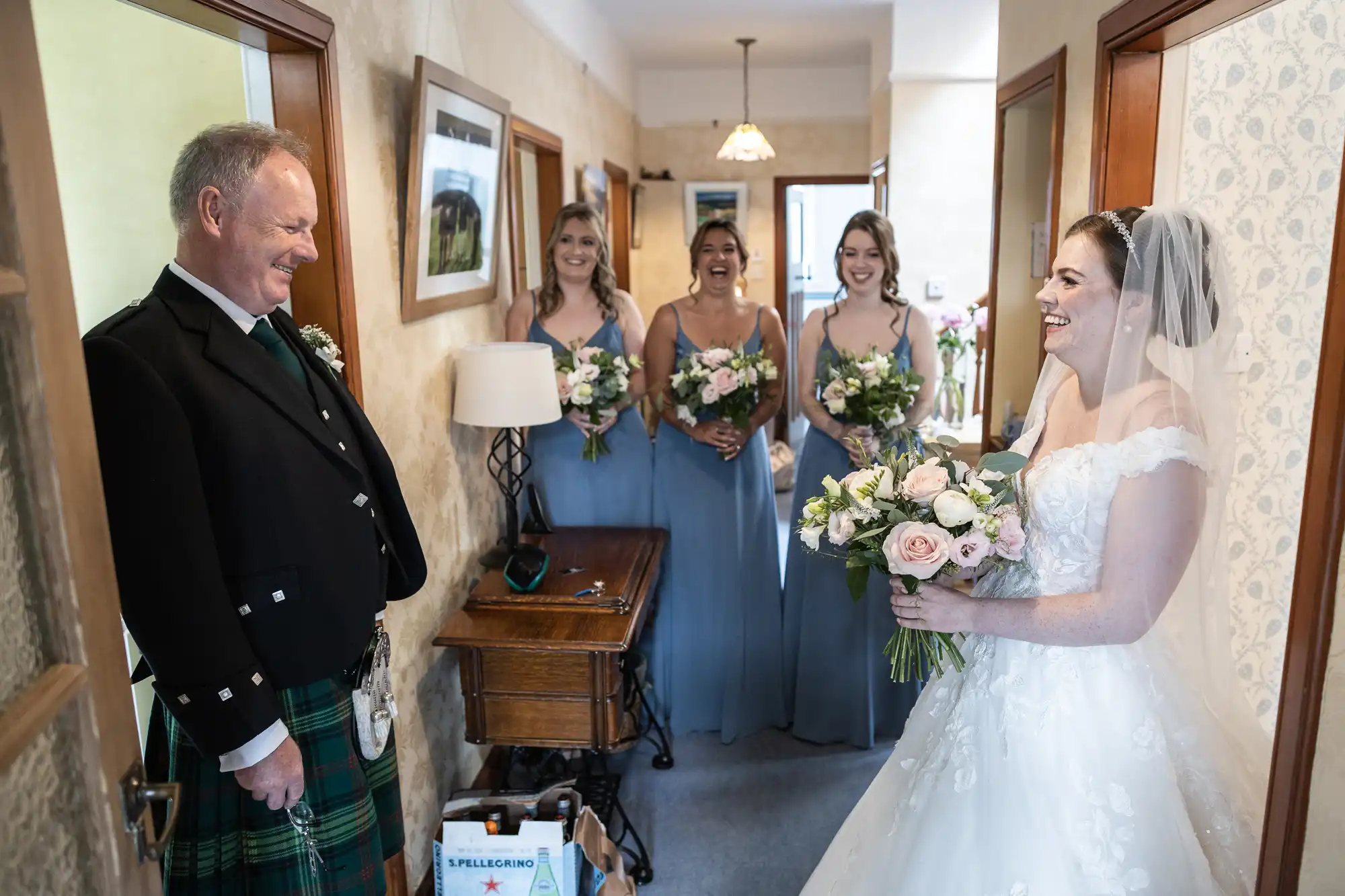 A bride in a white dress, holding a bouquet, smiles at an older man in a kilt. Three bridesmaids in light blue dresses with bouquets stand behind her in a room with beige walls and framed pictures.