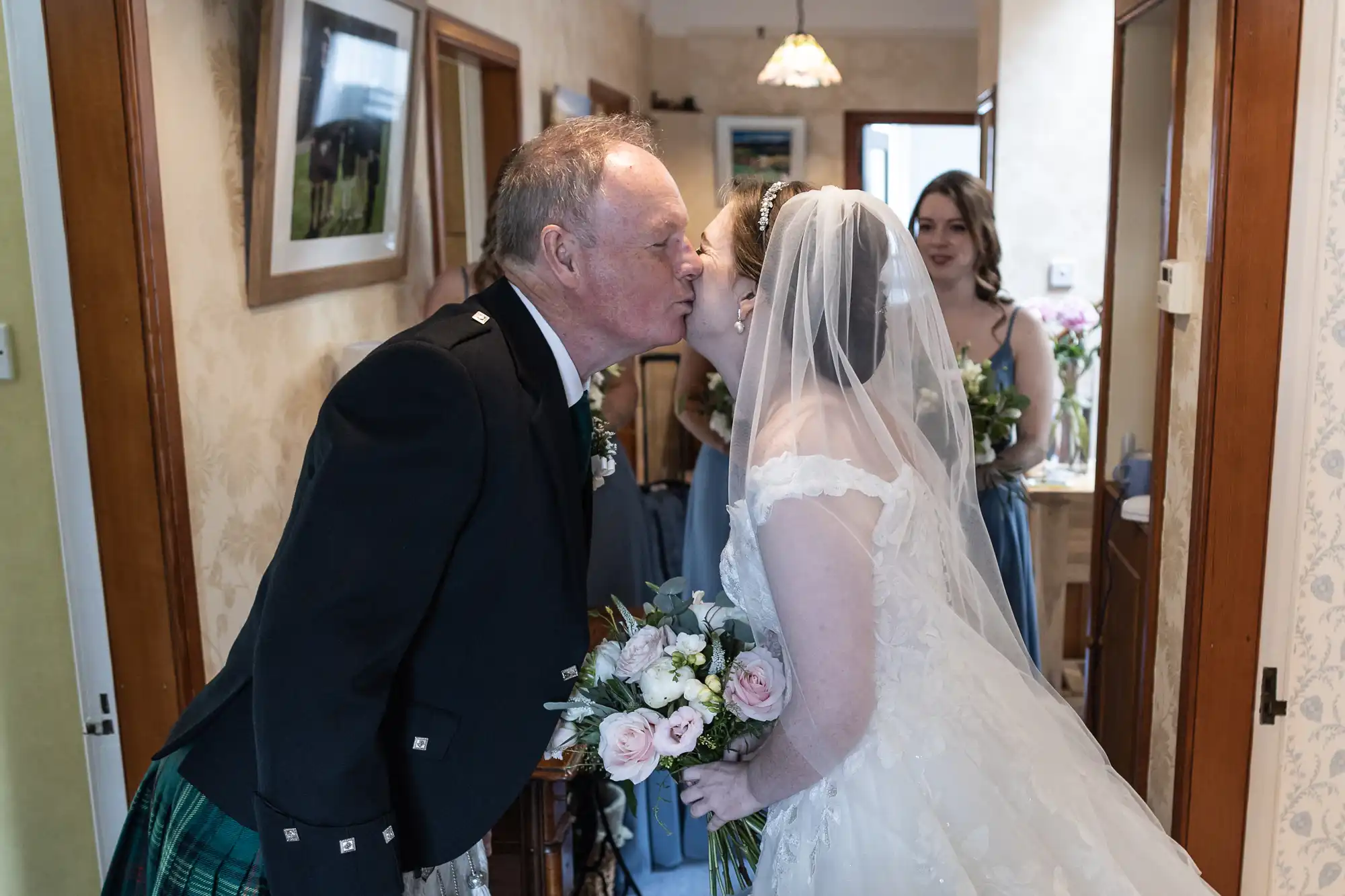 A bride in a white gown and veil kisses an older man in a suit in a hallway, with bridesmaids holding bouquets in the background.