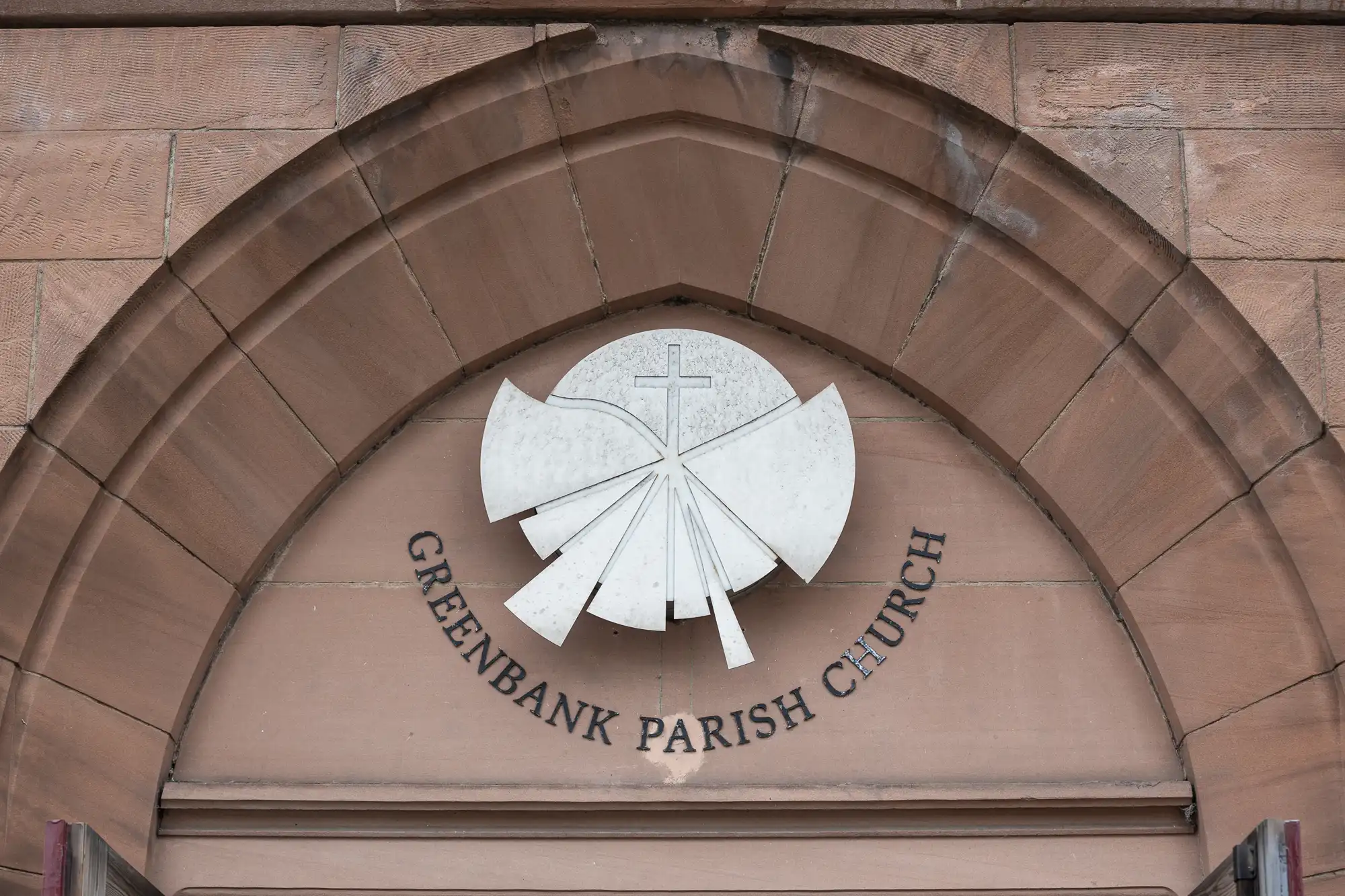 A stone arch displays a circular white emblem with a cross and rays, above the text "Greenbank Parish Church.