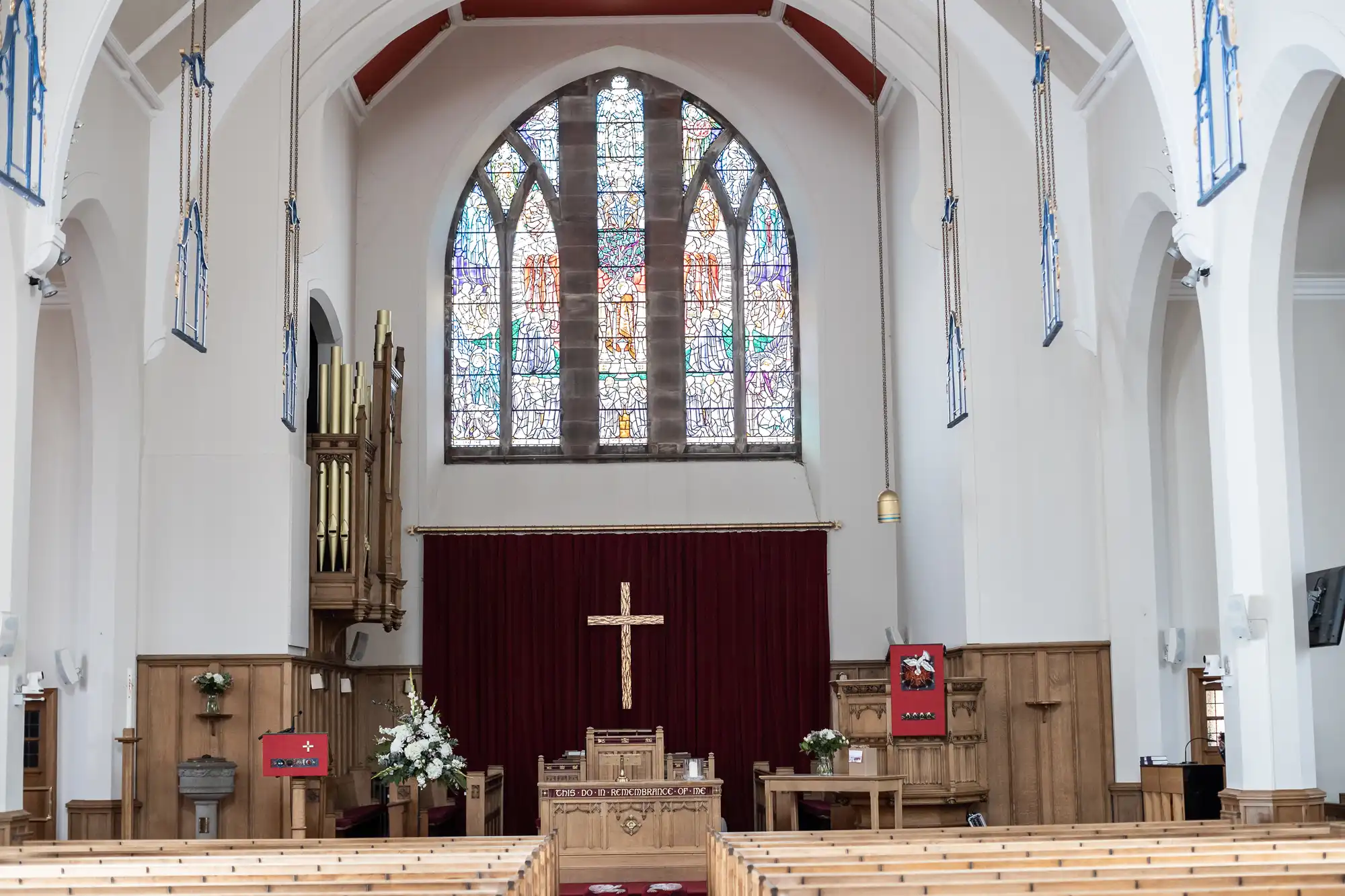 Interior of a church featuring wooden pews, a large stained-glass window, a central altar with a cross, and an organ to the left. Red banners and floral arrangements are present near the altar.