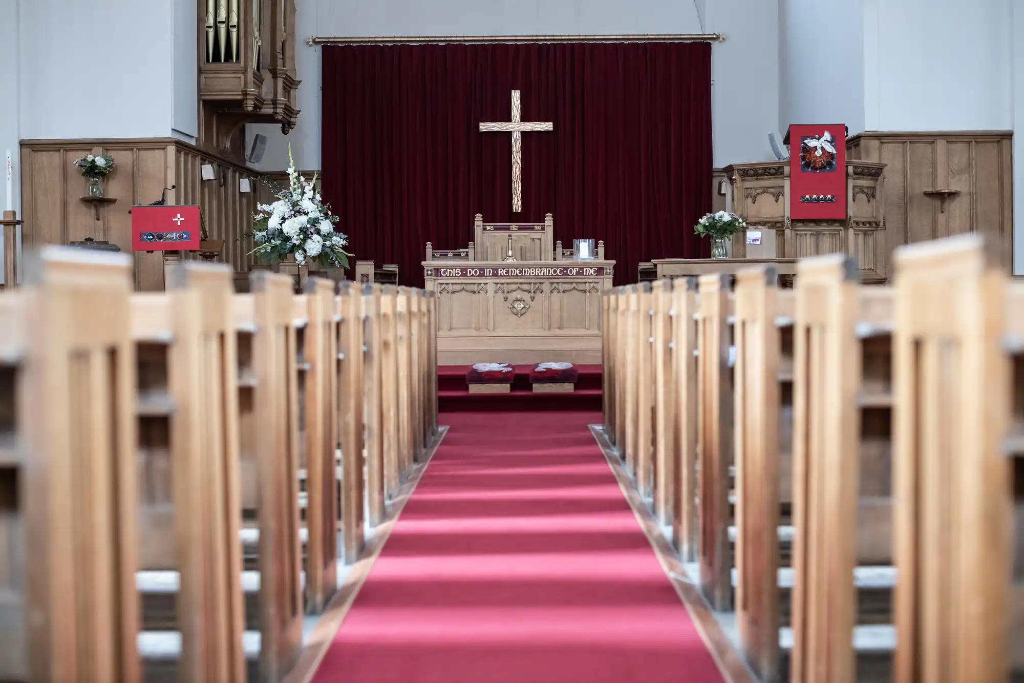 Interior of a church with wooden pews, a red carpeted aisle leading to an altar adorned with a cross, flower arrangements, and lit candles.