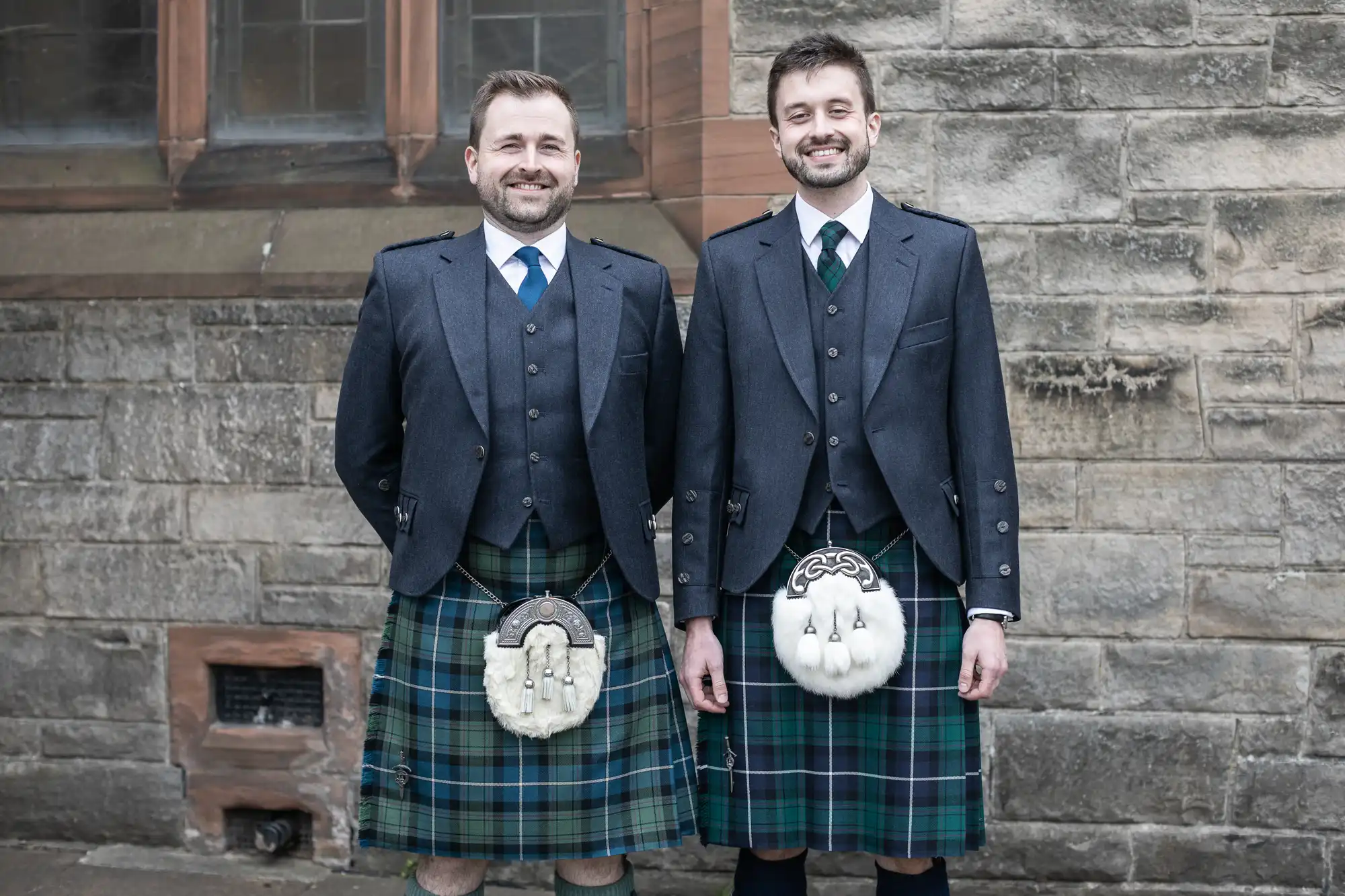 Two men smiling and standing side by side, wearing traditional Scottish kilts and jackets, in front of a stone building.