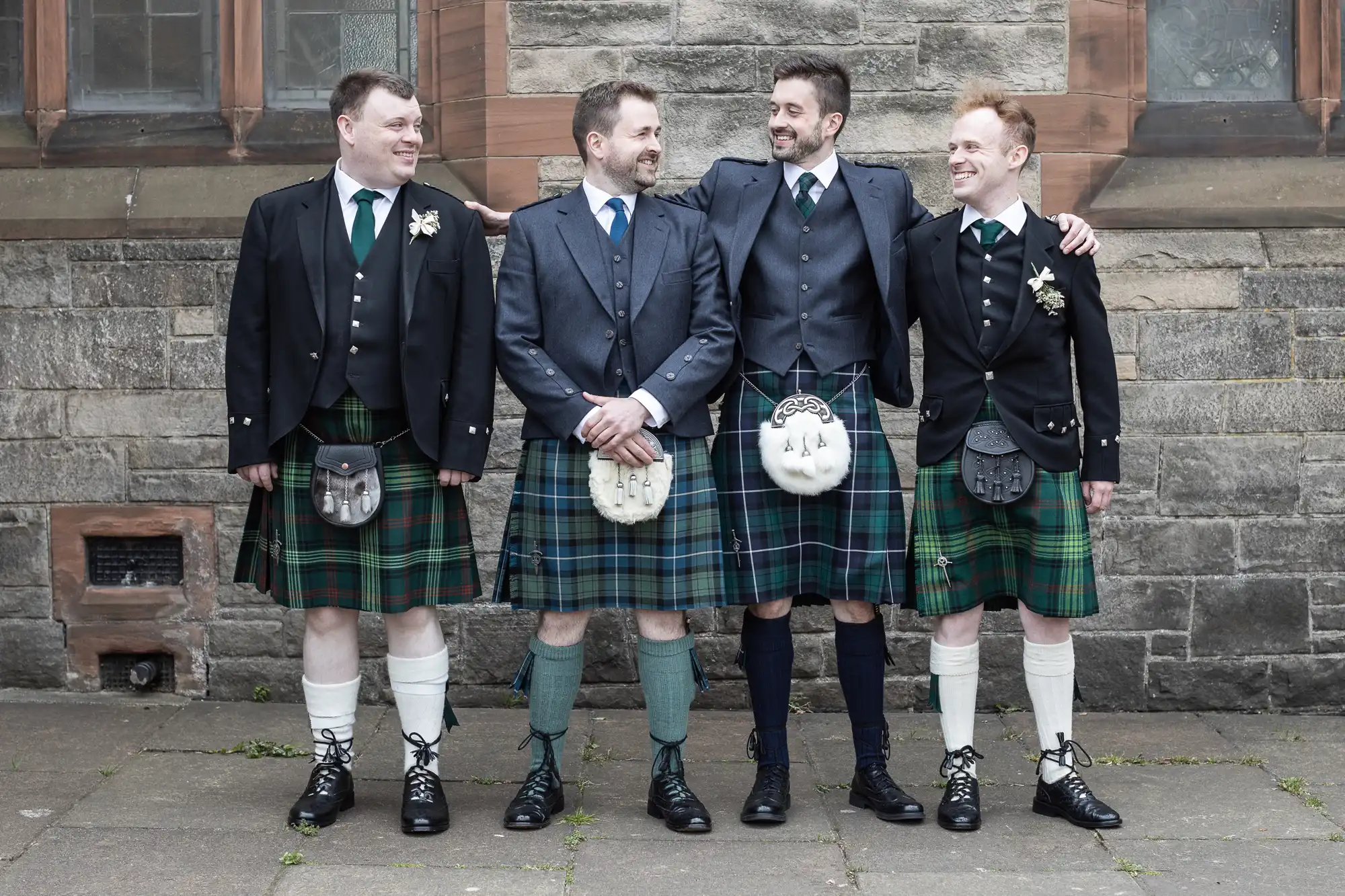 Four men in kilts stand in front of a stone building, smiling and looking at each other.