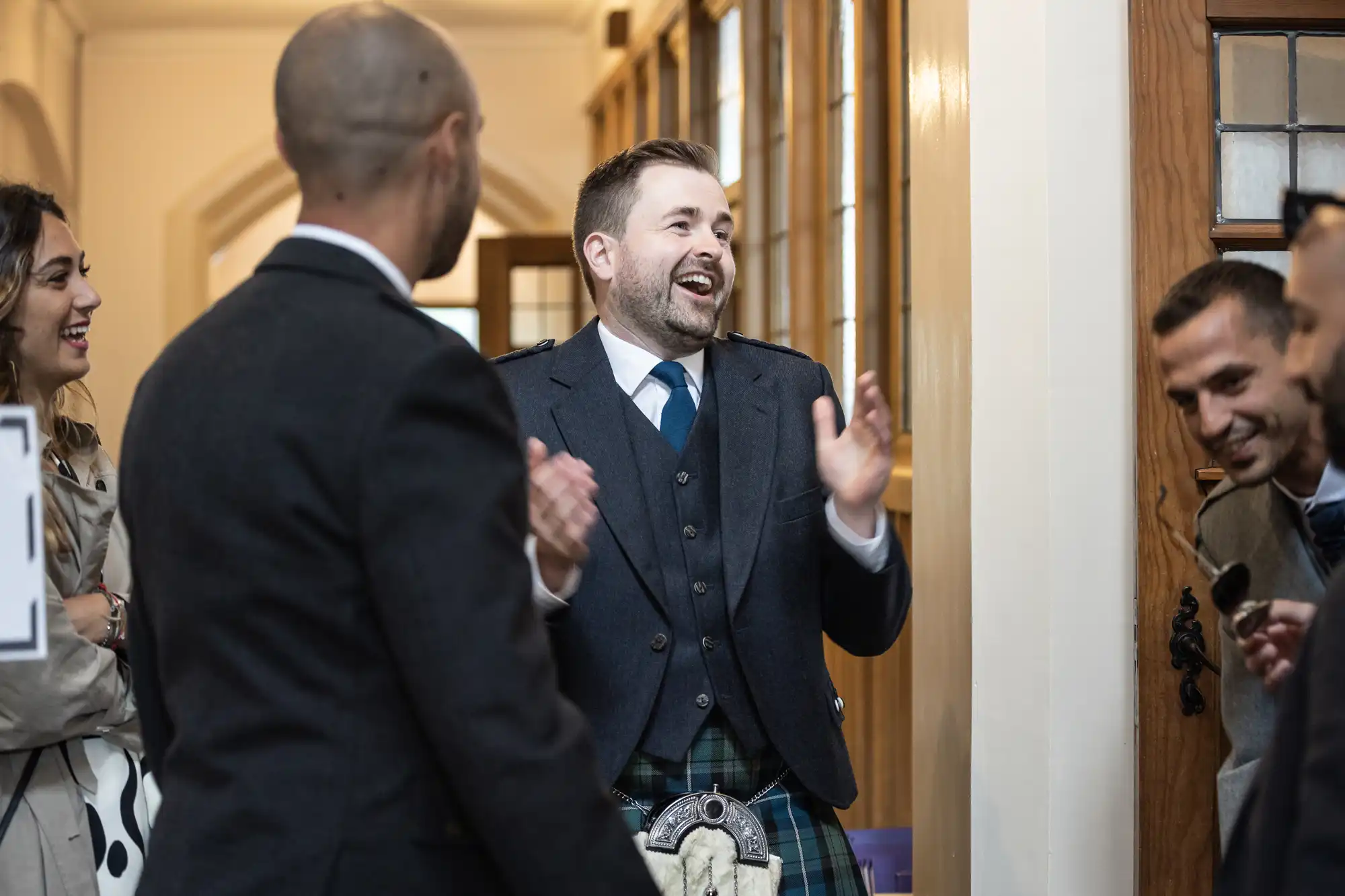 A man in a suit and tartan kilt claps and laughs with a group of people inside a hallway.