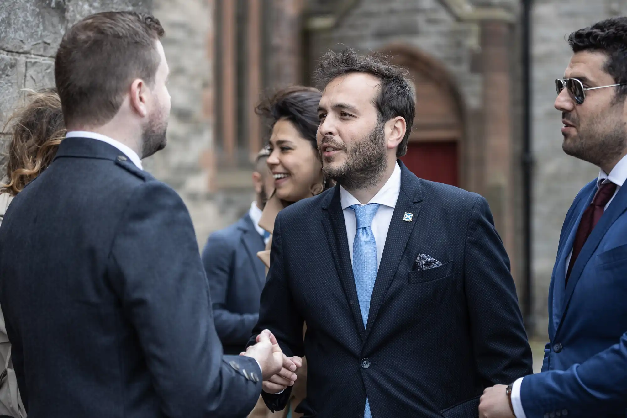 A group of people in formal attire are seen shaking hands and conversing outdoors near a stone building.