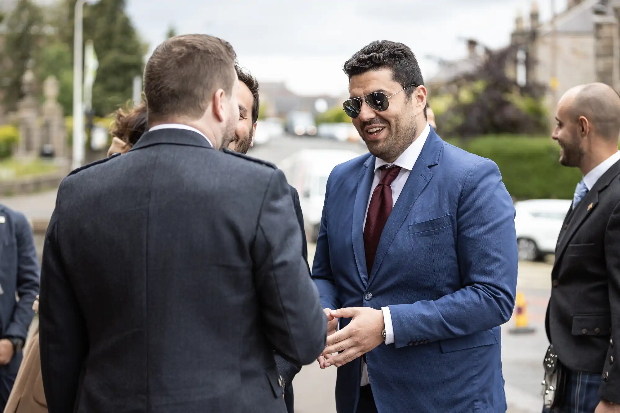 Four individuals in formal attire are engaged in conversation outdoors. One man in a blue suit and sunglasses is smiling, while another in a dark suit has his back to the camera.