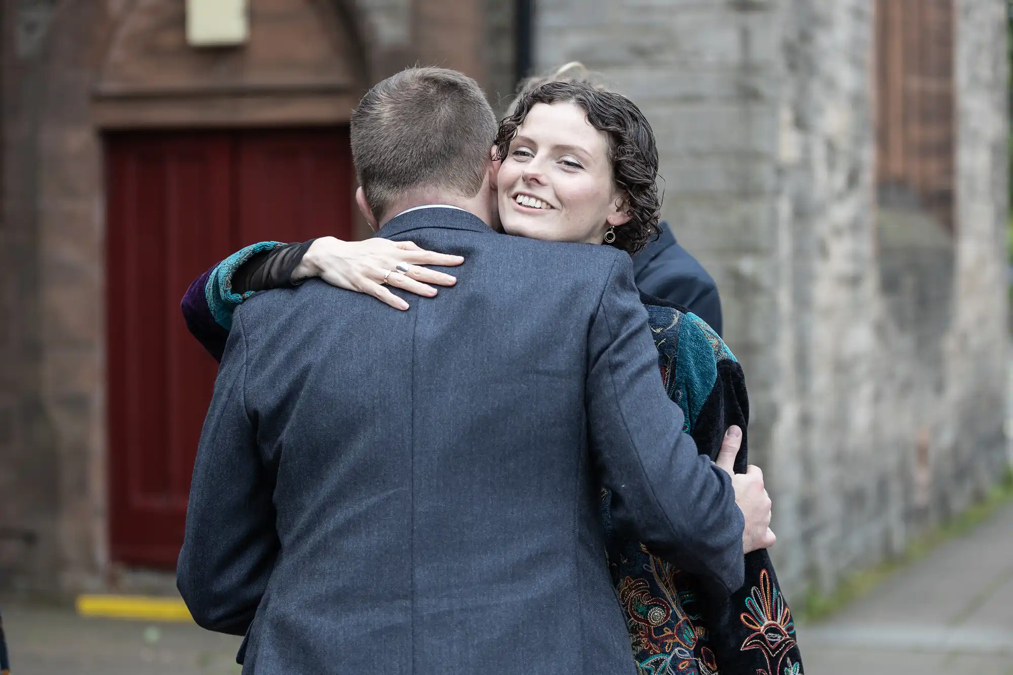 Two people embracing outdoors. The woman faces the camera, smiling, while the man has his back to the camera. They are in front of a stone building with a red door.