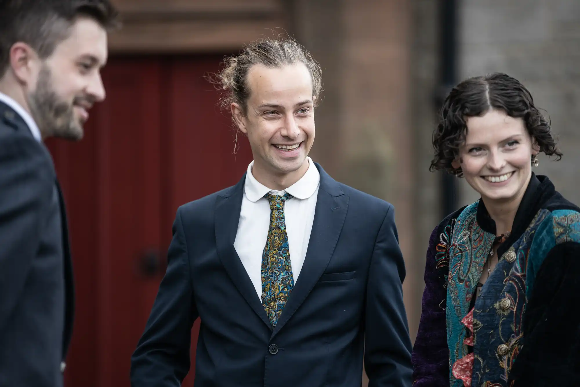 Three people in formal attire smile and converse outdoors. The two men wear suits, and the woman wears a multicolored jacket.