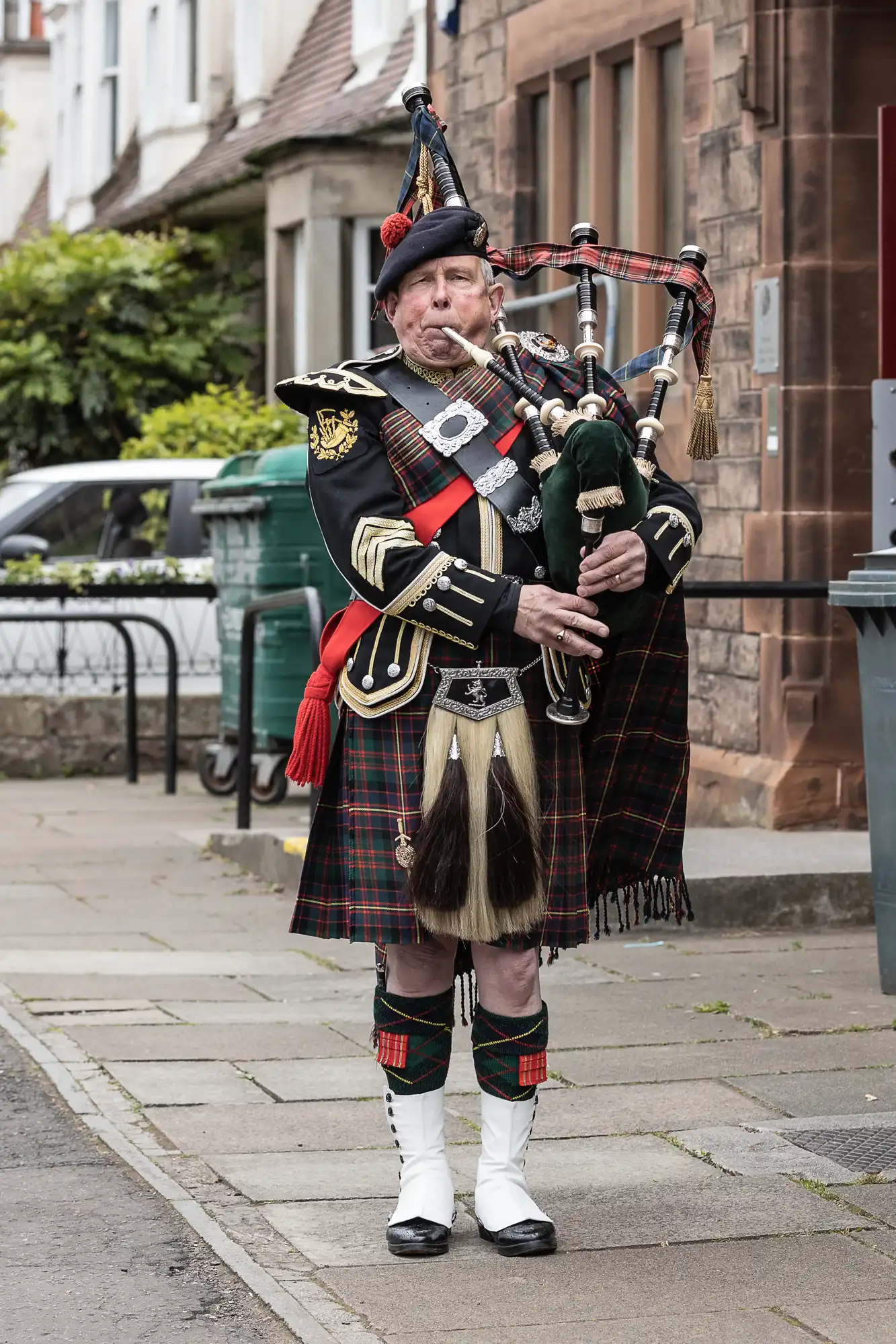 A man wearing a traditional Scottish kilt and uniform plays the bagpipes on a sidewalk. He stands in front of a stone building and paved walkway.