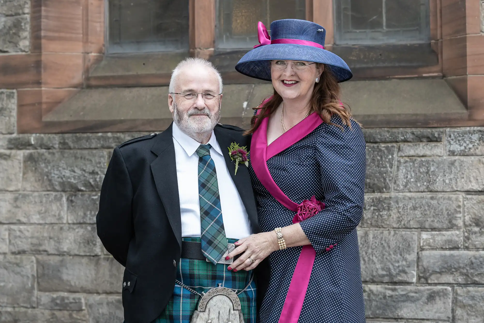 A man and woman pose in front of a stone building, with the man wearing a traditional Scottish kilt and the woman wearing a polka dot dress and large blue hat.