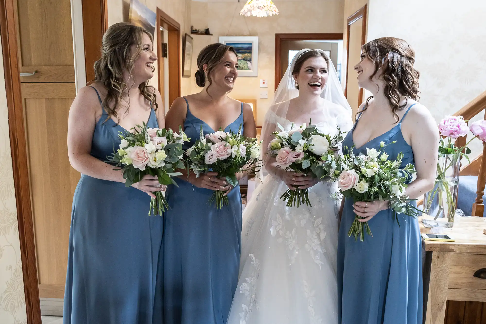 A bride in a white gown stands with three bridesmaids in blue dresses, all holding floral bouquets. They are indoors, smiling, and conversing.