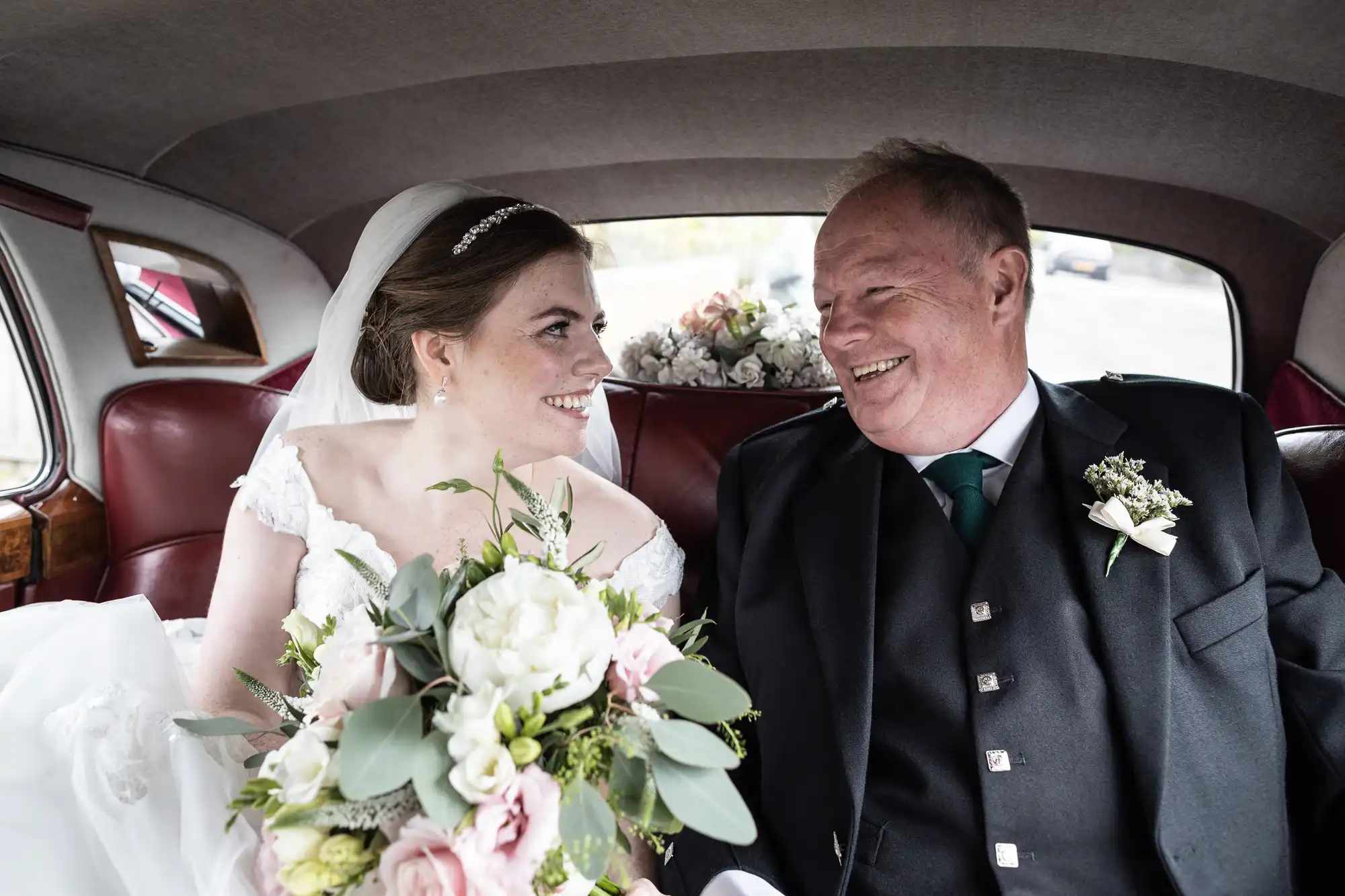 A bride holding a bouquet and an older man, both well-dressed and smiling, sit together in the backseat of a car adorned with flowers.