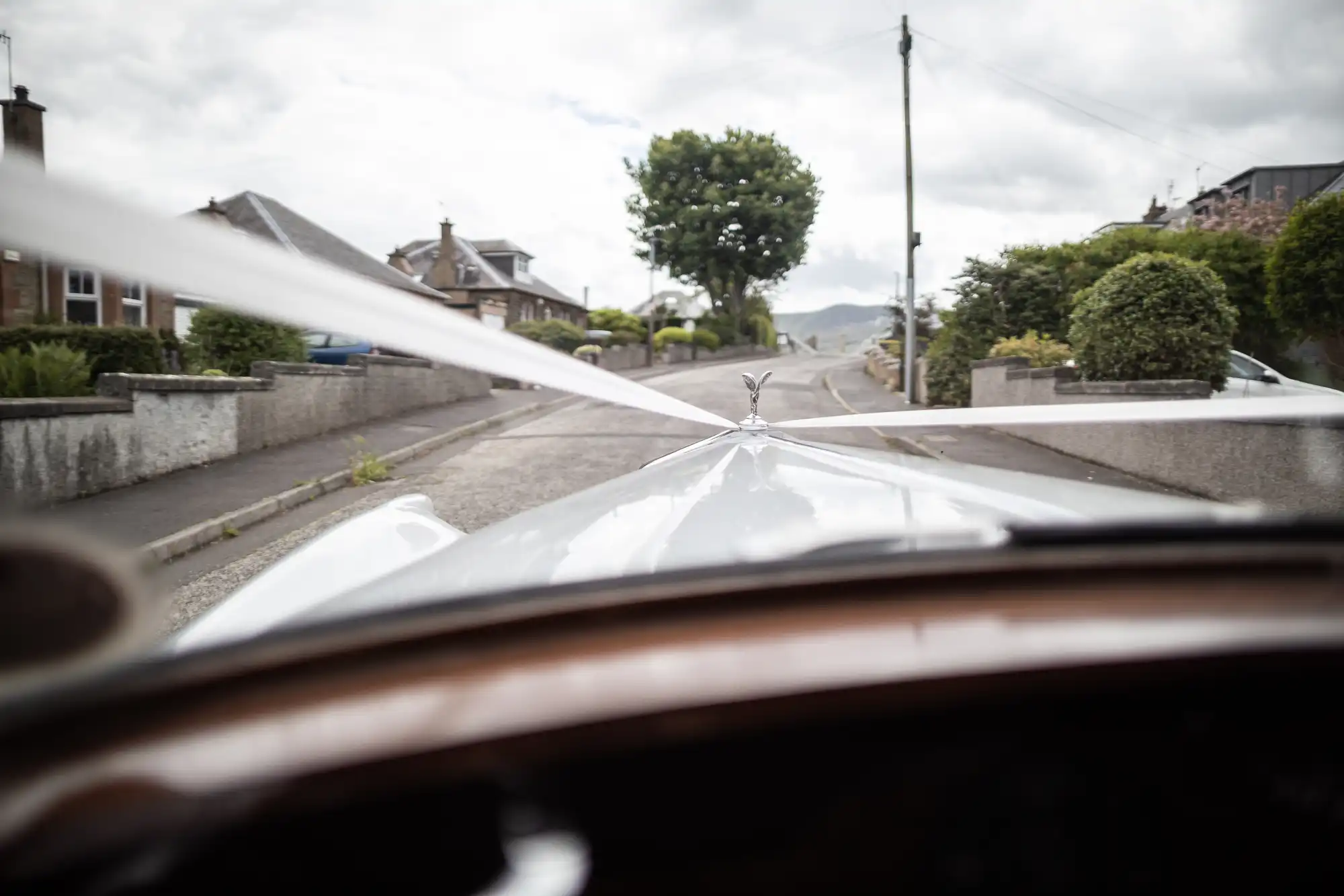 View from a car's windshield decorated with white ribbon driving down a suburban street with houses and trees under a cloudy sky.