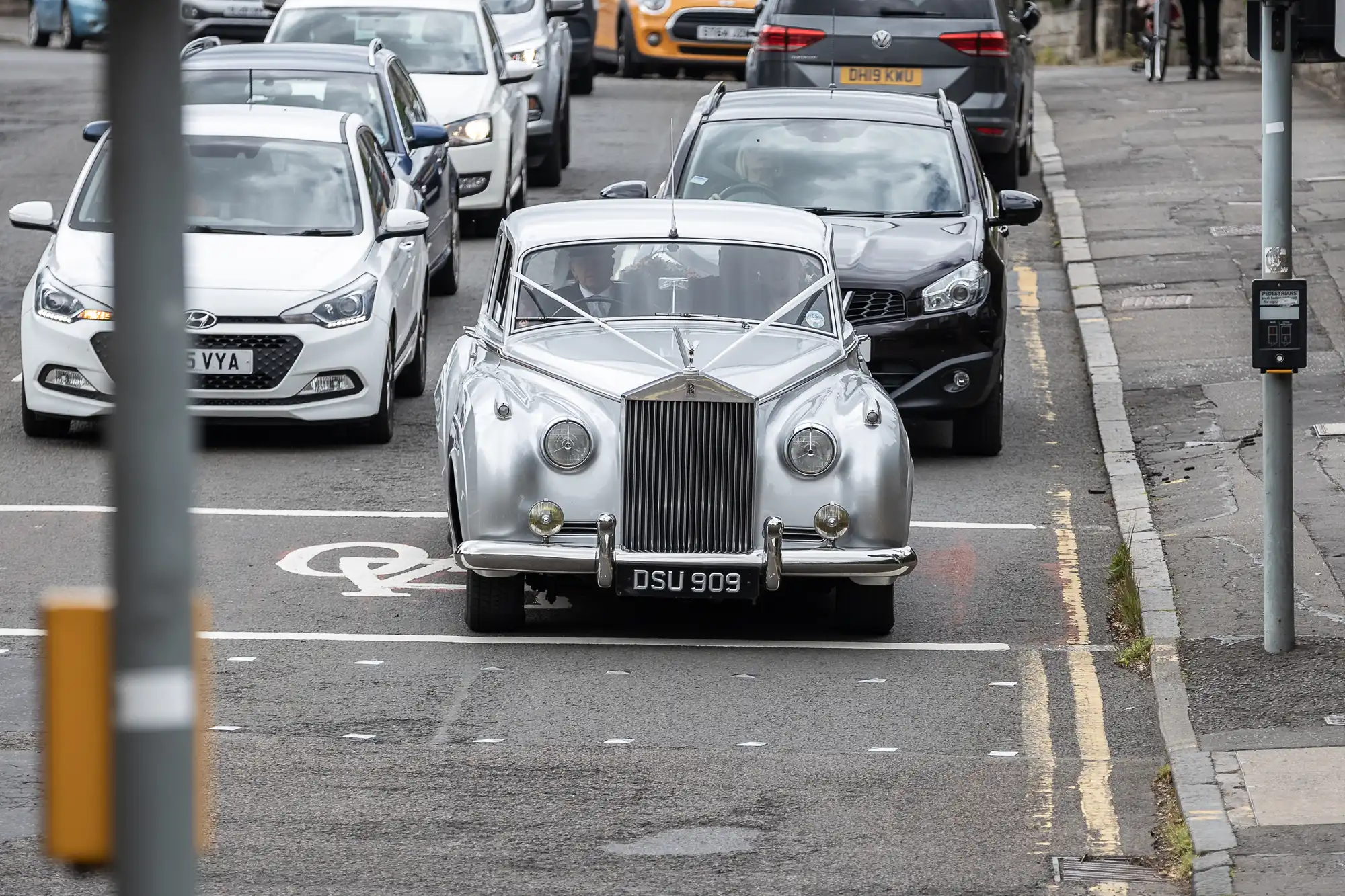 A vintage silver car is stopped at a traffic light, surrounded by modern vehicles on a city street.