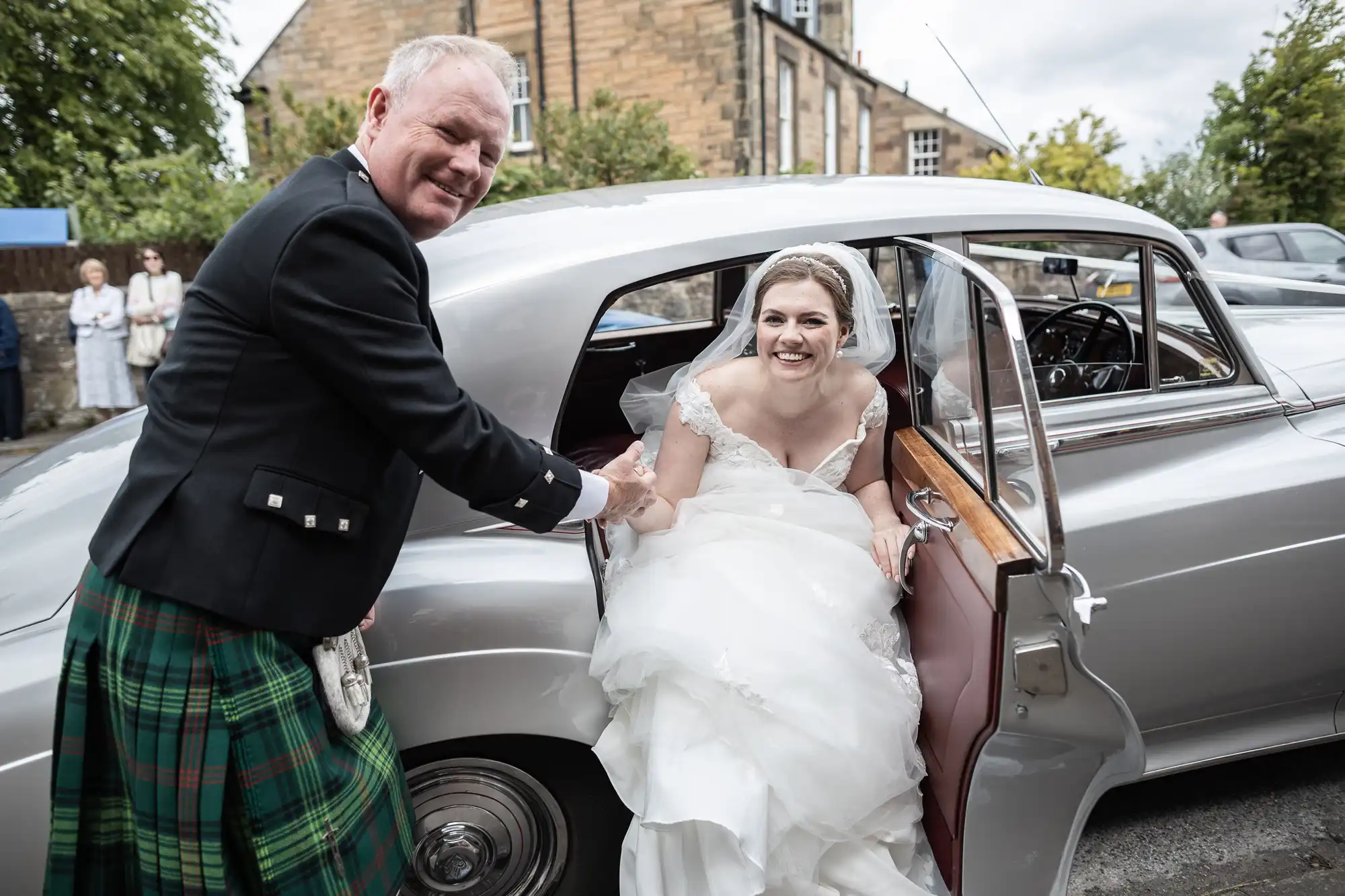 A man in a kilt helps a smiling bride exit a vintage car outside a stone building.