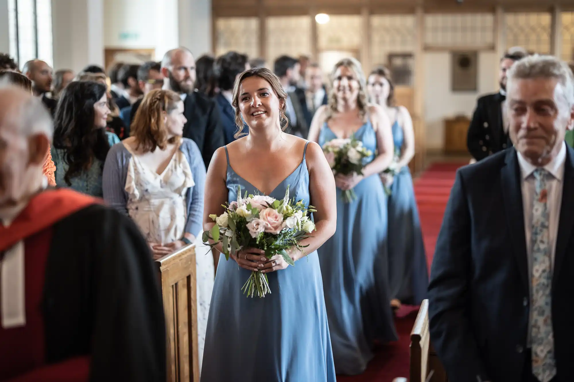 A group of women in blue dresses, holding bouquets, walk down the aisle of a church. Other people are seated and standing, watching the procession. The interior of the church is visible in the background.