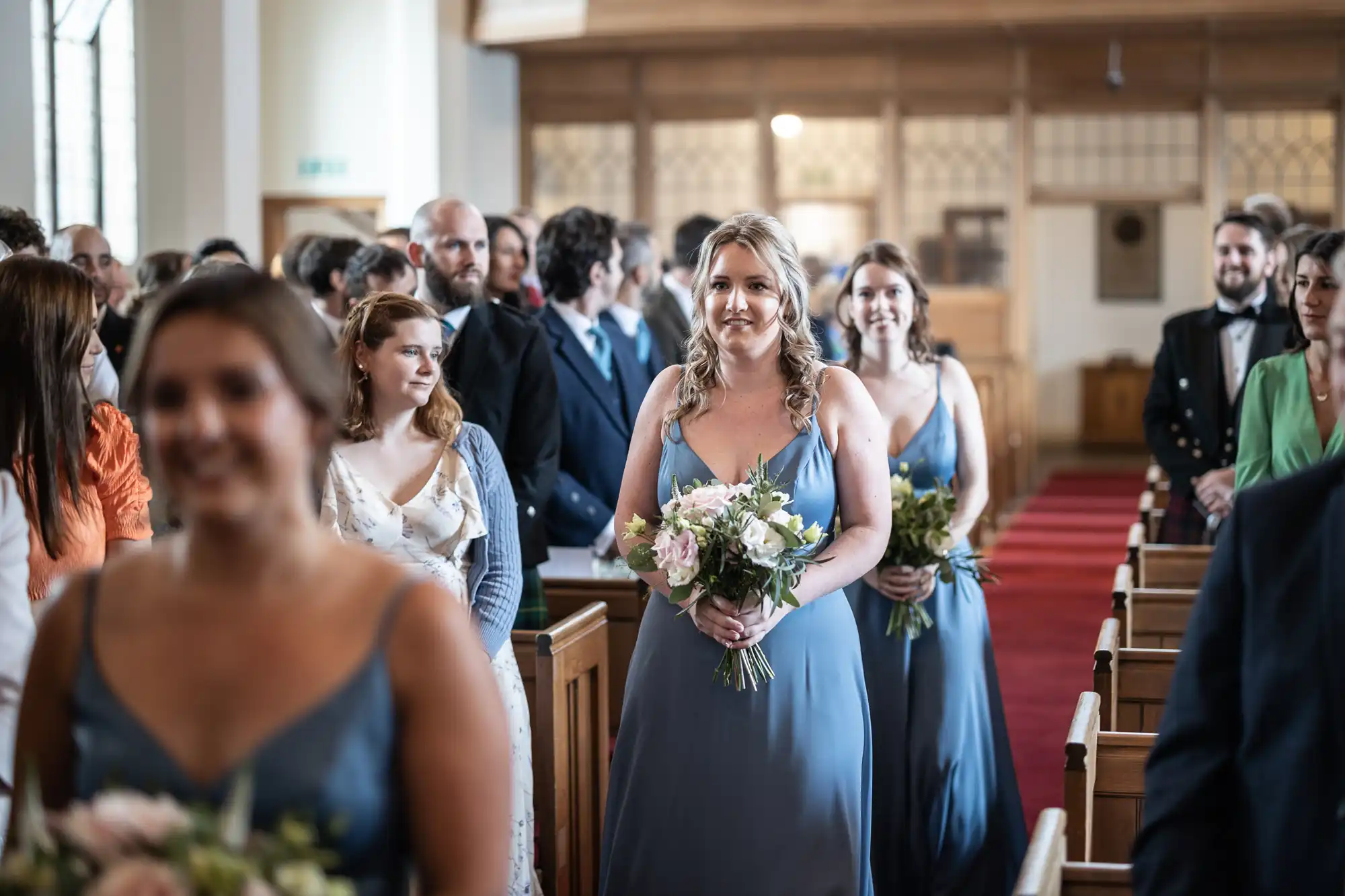 Bridesmaids in blue dresses hold bouquets as they walk down the aisle of a church, with guests seated on either side.