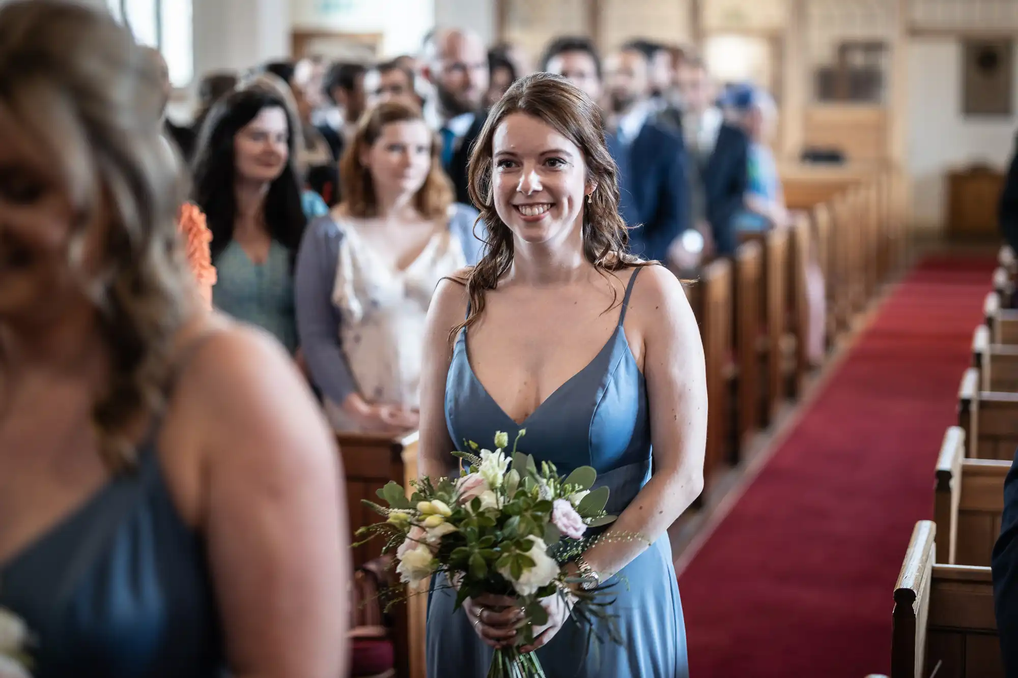 A woman in a blue dress holding a bouquet of flowers smiles while walking down the aisle of a church, with seated guests in the background.