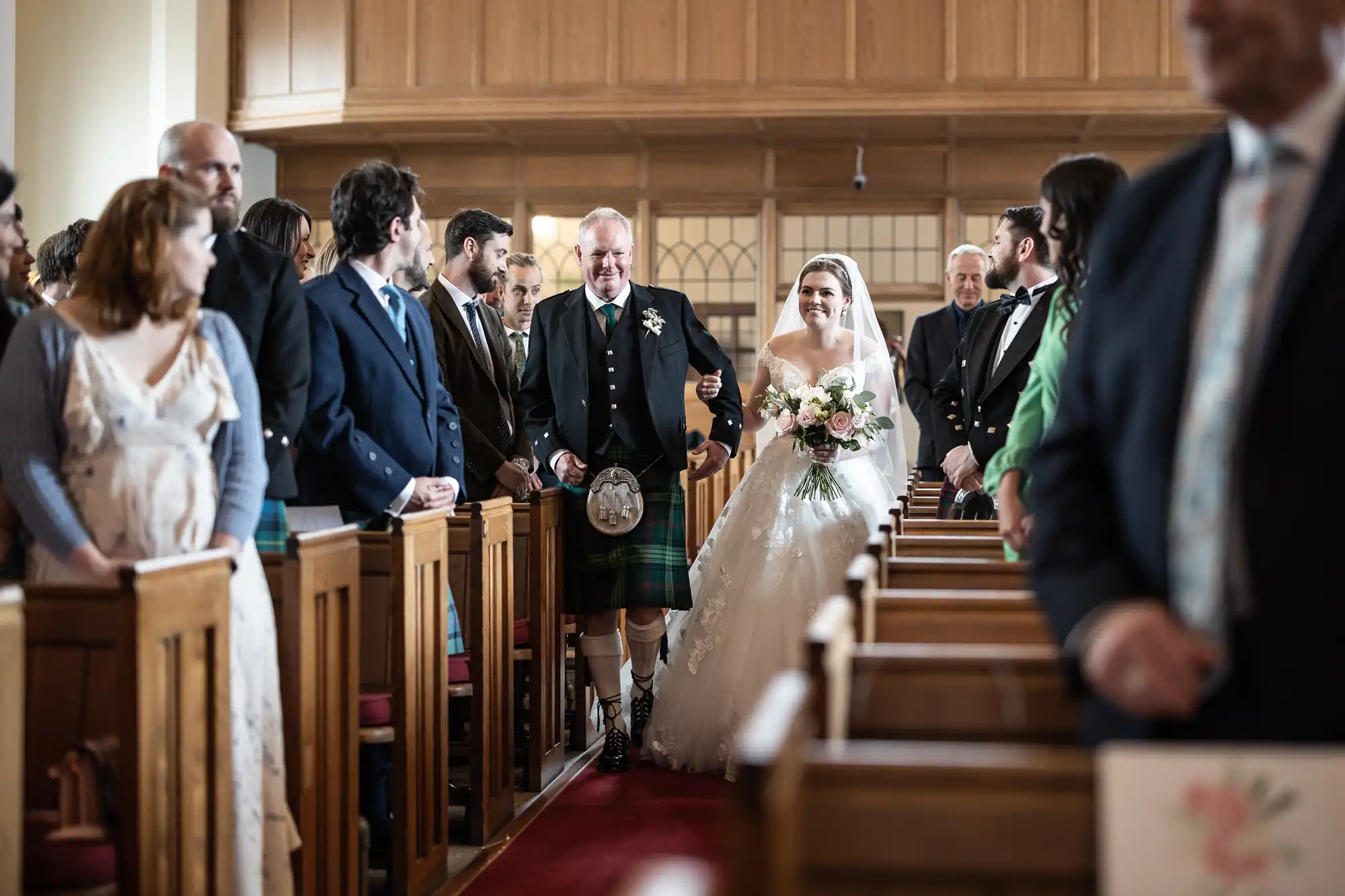 A bride, escorted by a man in a kilt, walks down the aisle of a church, surrounded by standing guests.