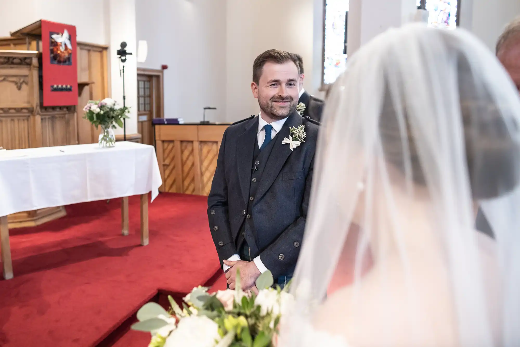 A groom in a dark suit looks at the bride in a wedding dress and veil inside a church with red carpet and wooden furnishings.