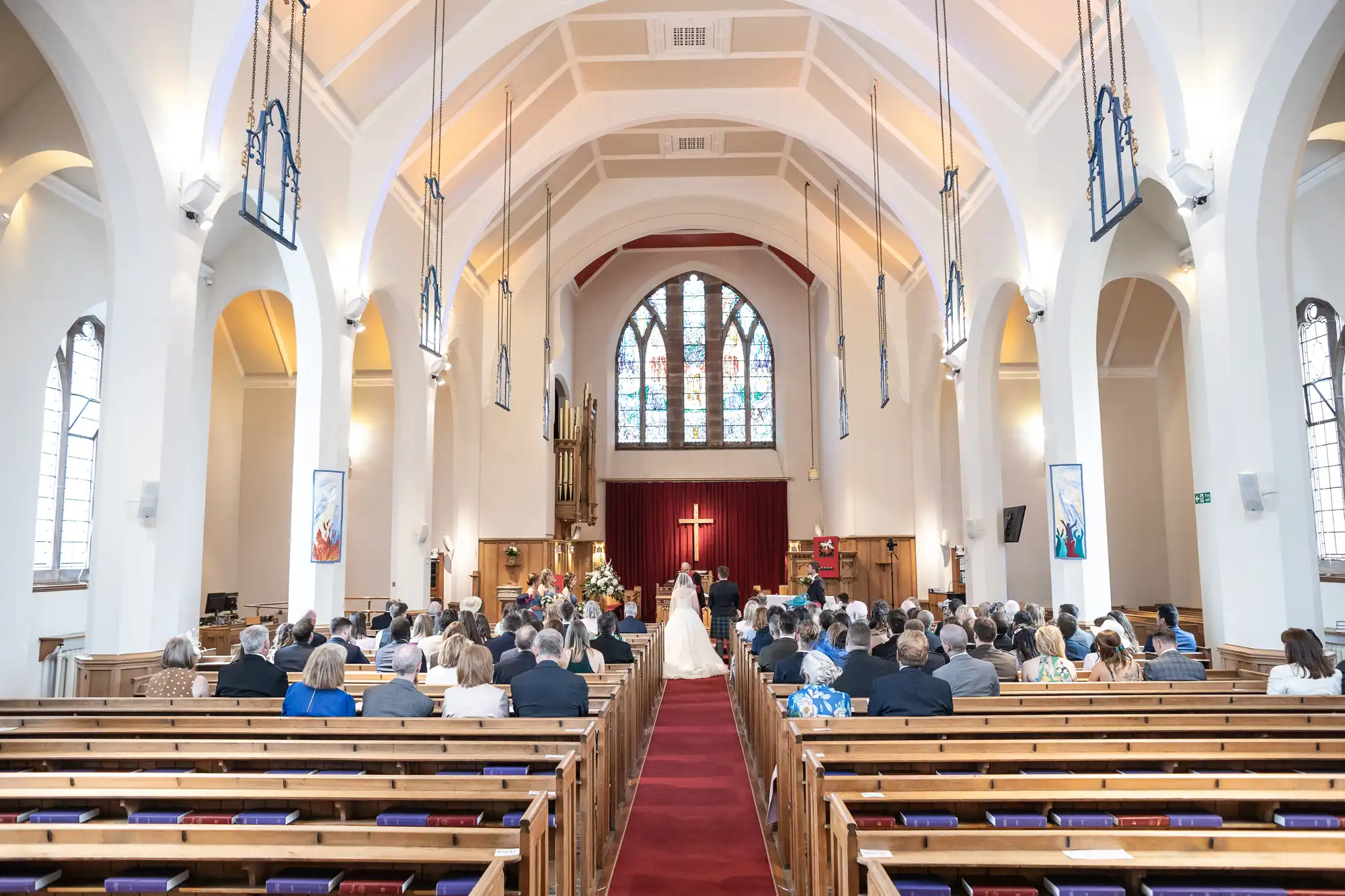 A bride and groom stand at the altar in front of an audience in a large church with a high arched ceiling, stained glass windows, and a red carpet aisle.