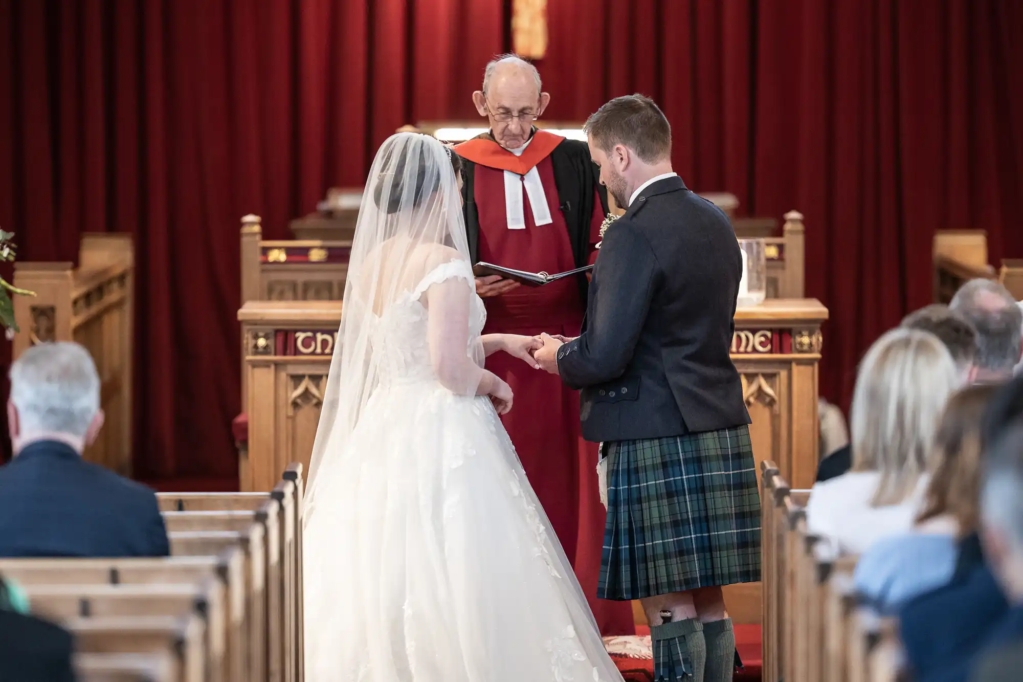 A bride and groom stand before a minister during their wedding ceremony in a church, with the groom placing a ring on the bride's finger.