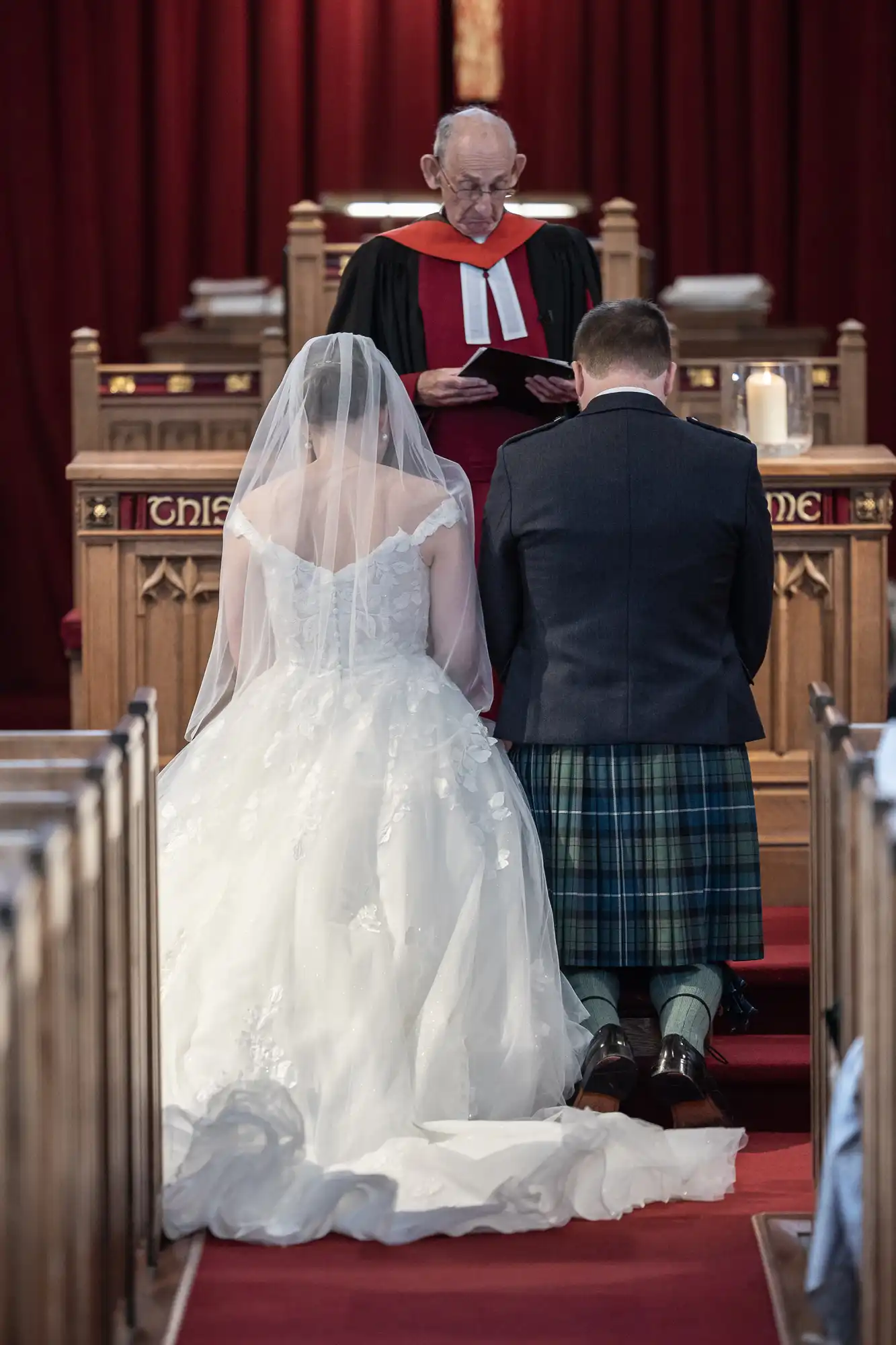 A bride and groom kneel before a priest in a church ceremony. The bride wears a white dress with a long veil, and the groom wears a kilt.