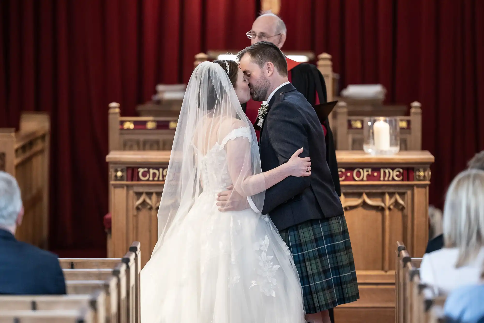 A bride in a white gown and a groom in a kilt share a kiss in a church in front of a clergy member during their wedding ceremony.