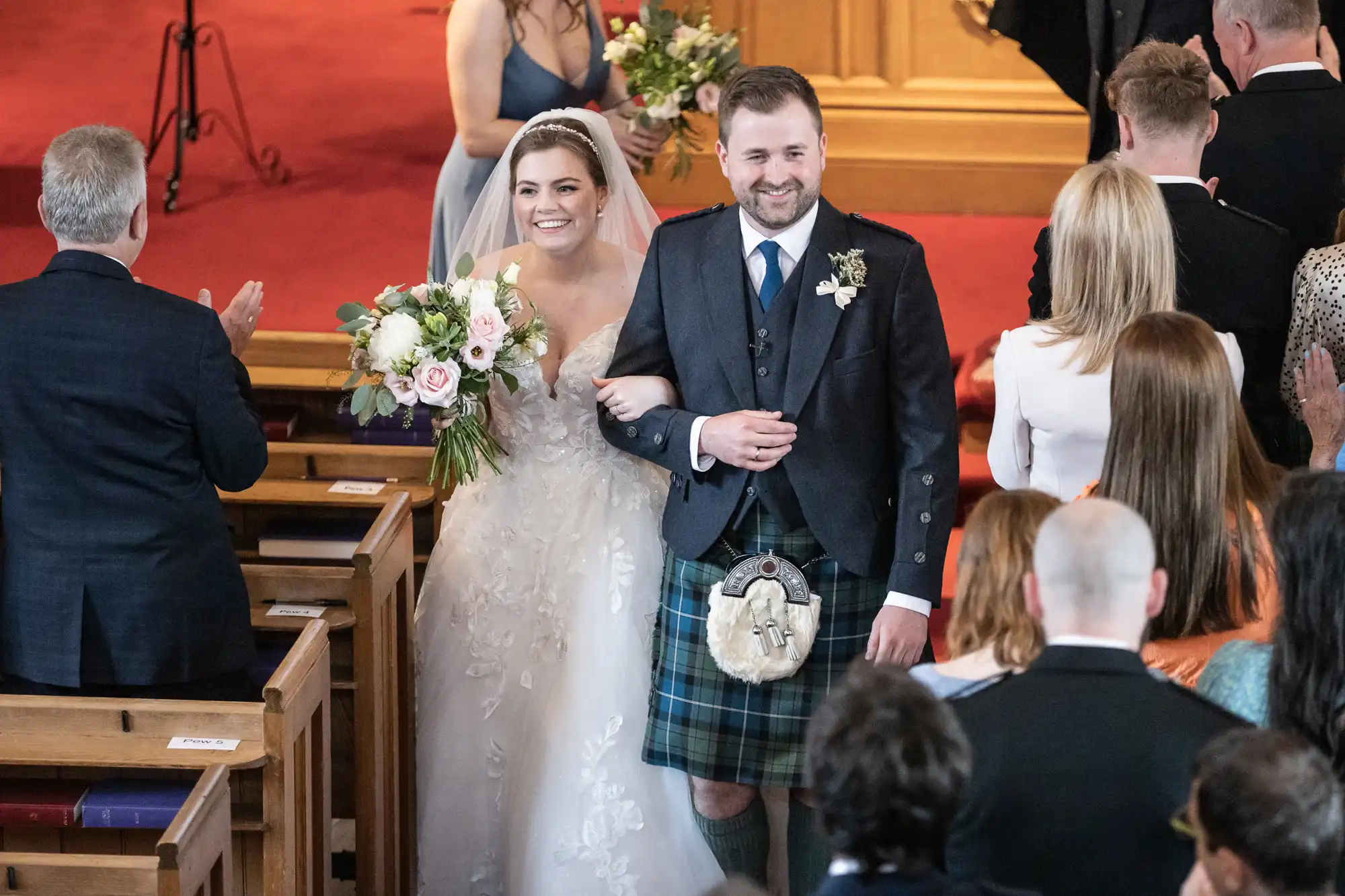 A bride and groom walk down the aisle together in a church, smiling while guests look on. The groom wears a kilt, and the bride holds a bouquet of flowers.