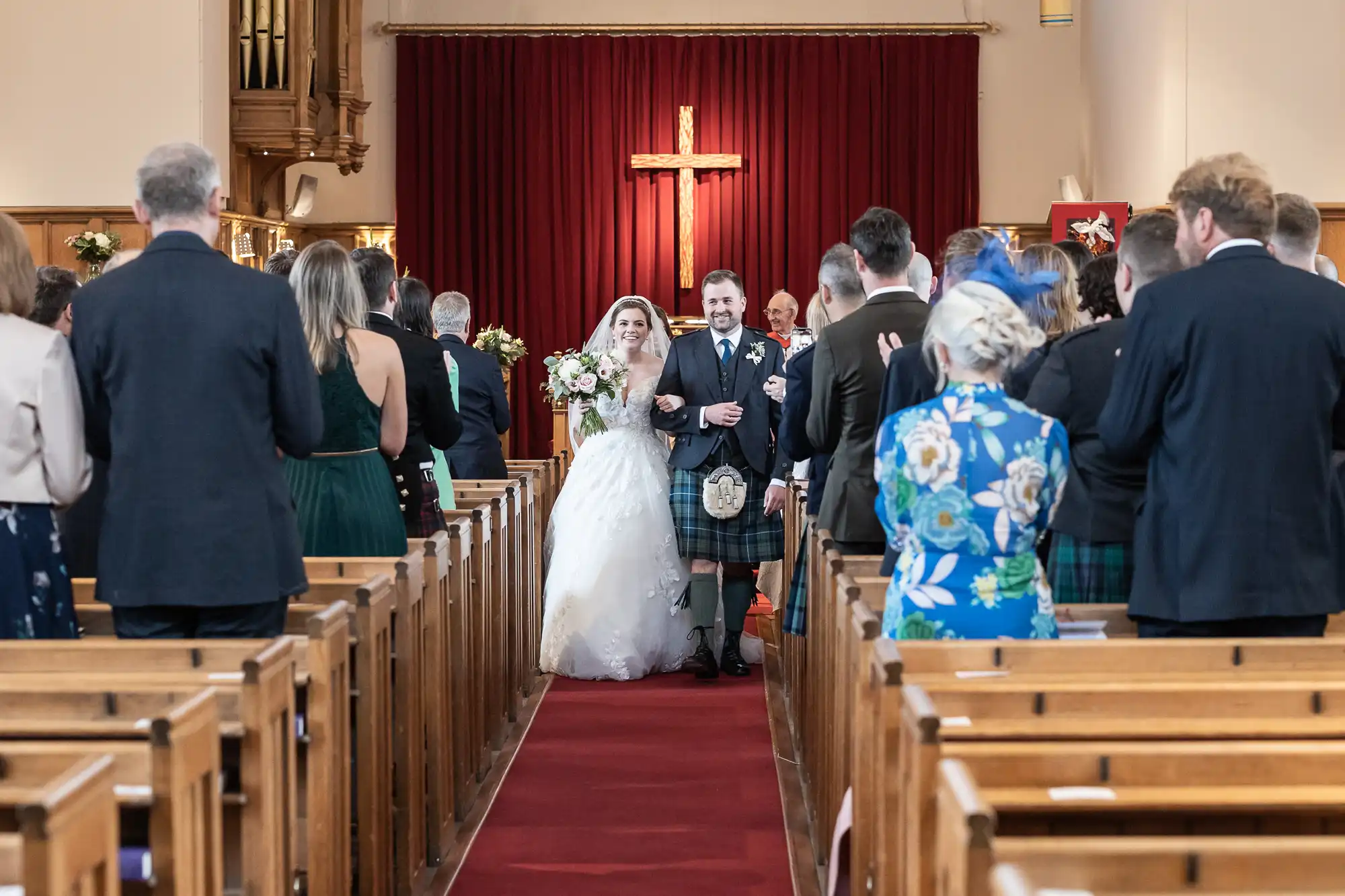 A bride and groom walk down the aisle together in a church. Guests stand on either side, observing the couple. The groom wears a kilt, and a large wooden cross is displayed at the front.