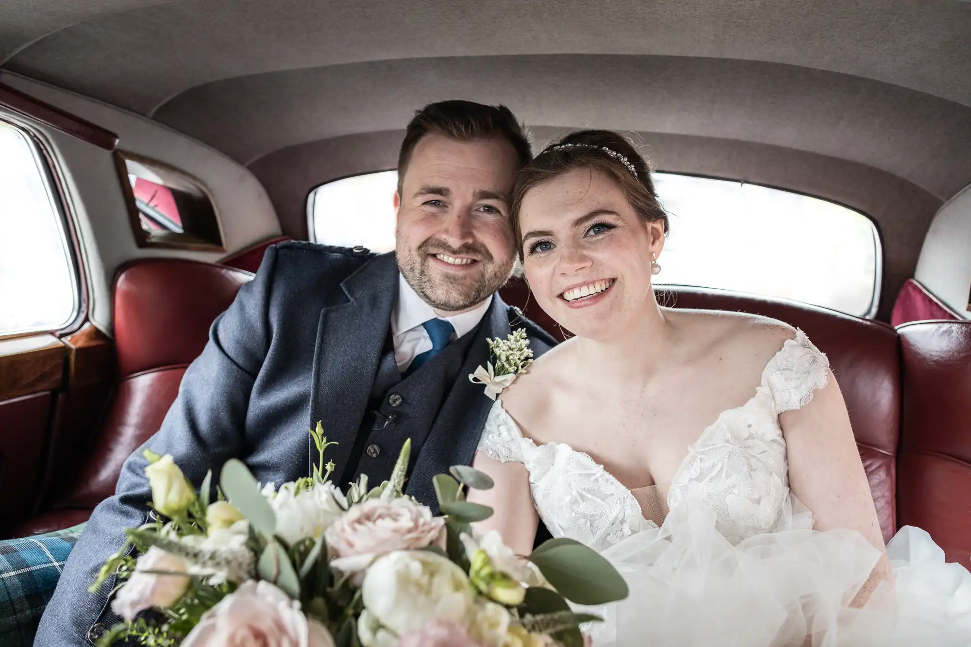 A smiling couple dressed in wedding attire sit together in the back of a car. The bride holds a bouquet of flowers.