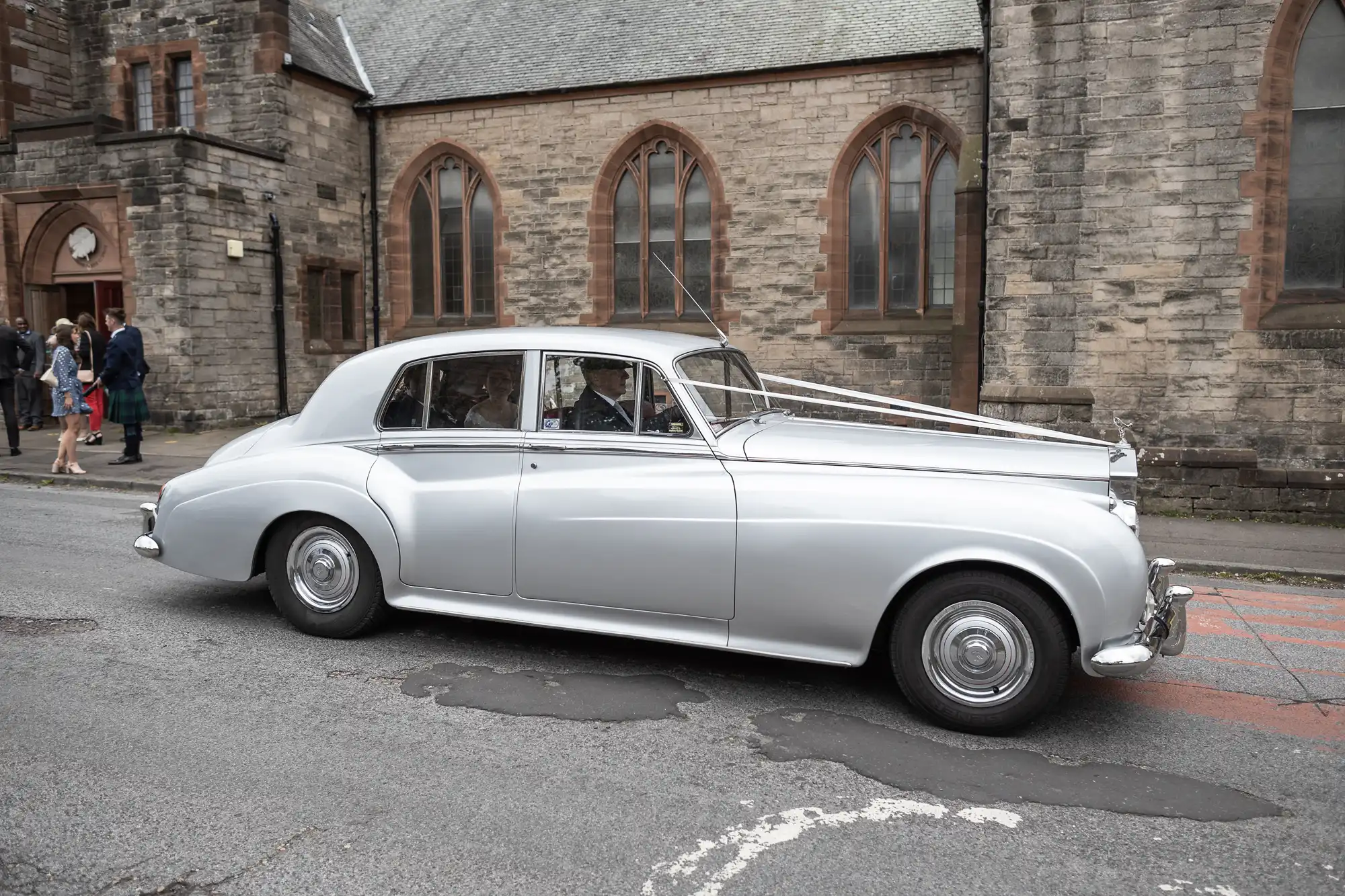 A silver classic car is parked on a street in front of a stone church. A few people are visible in the background, near the church entrance.