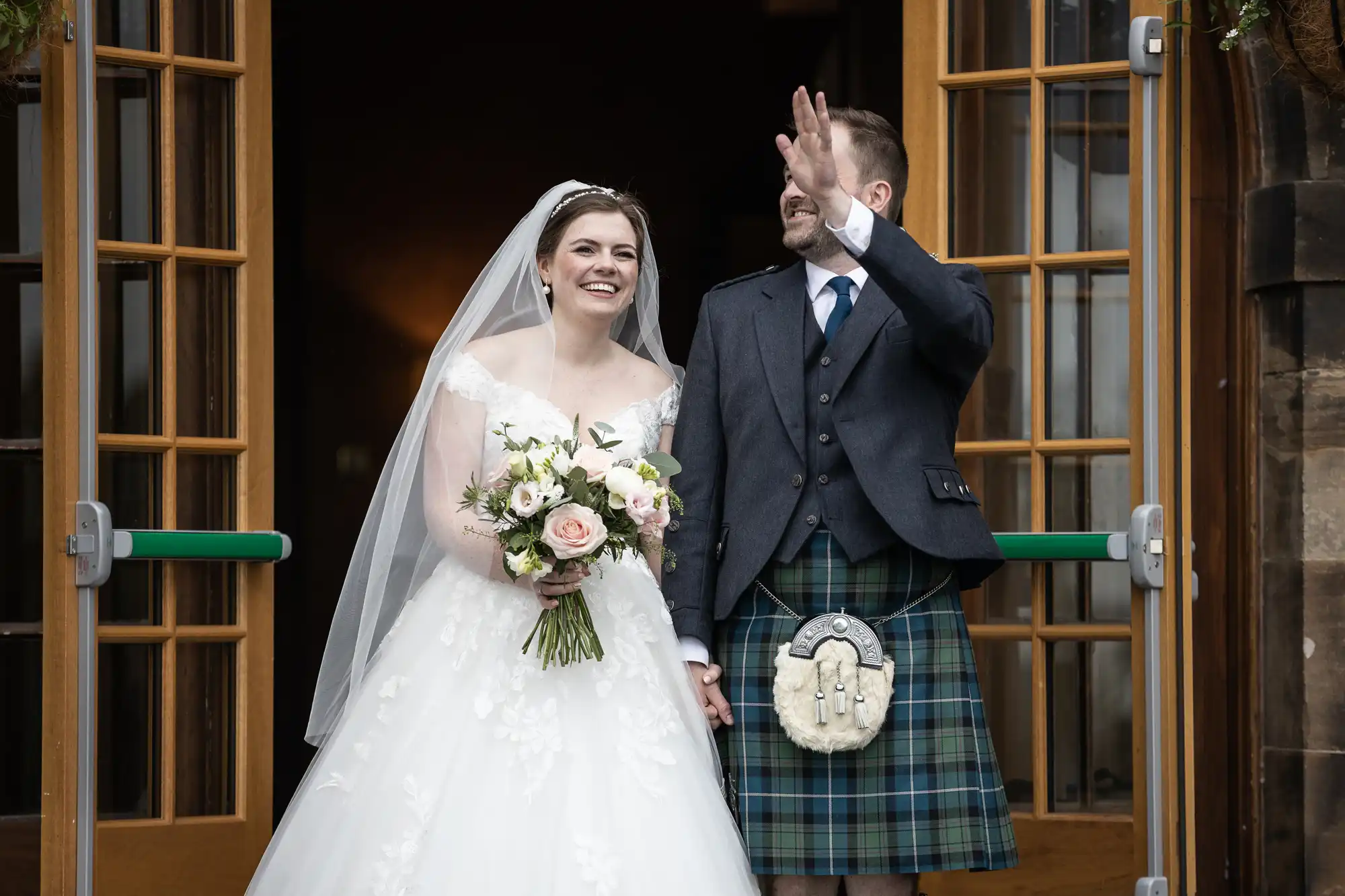 A bride in a white wedding dress and groom in a kilt stand at double doors; she holds a bouquet while he waves.