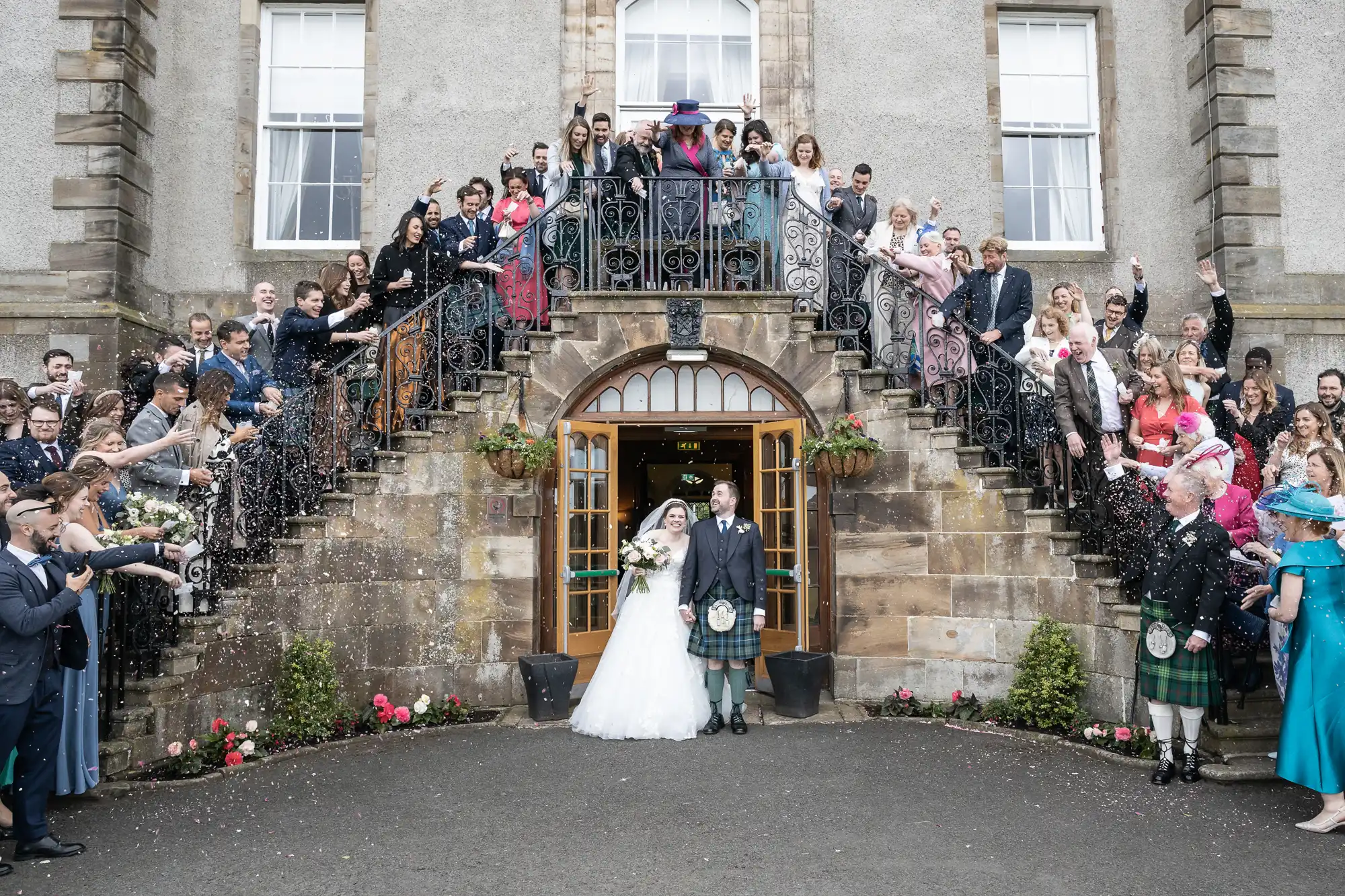 A newlywed couple stands in front of a grand building's entrance, surrounded by cheering guests on staircases, who are throwing confetti. The scene is lively and celebratory.