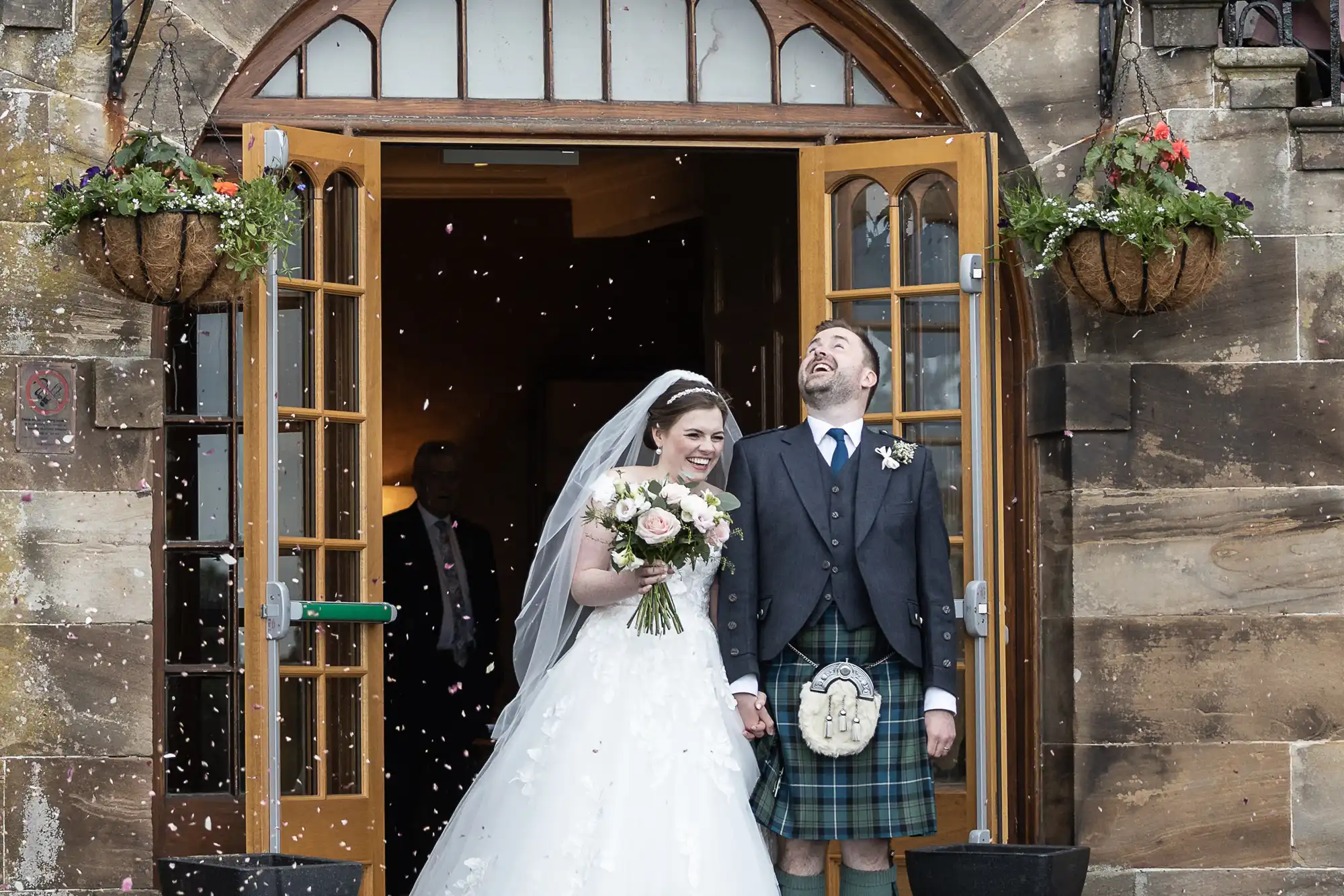 A couple in wedding attire, the bride in a white gown and the groom in a kilt, are standing at an open doorway, smiling and laughing as flower petals are thrown.