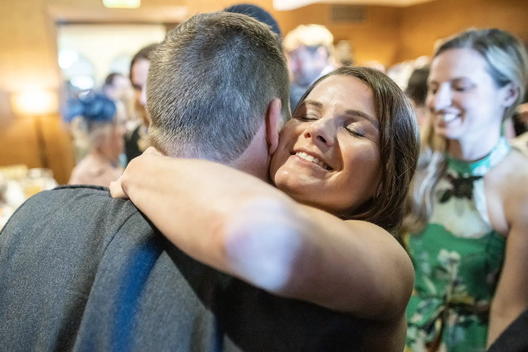 A woman embraces a man while smiling, in a room filled with other people.