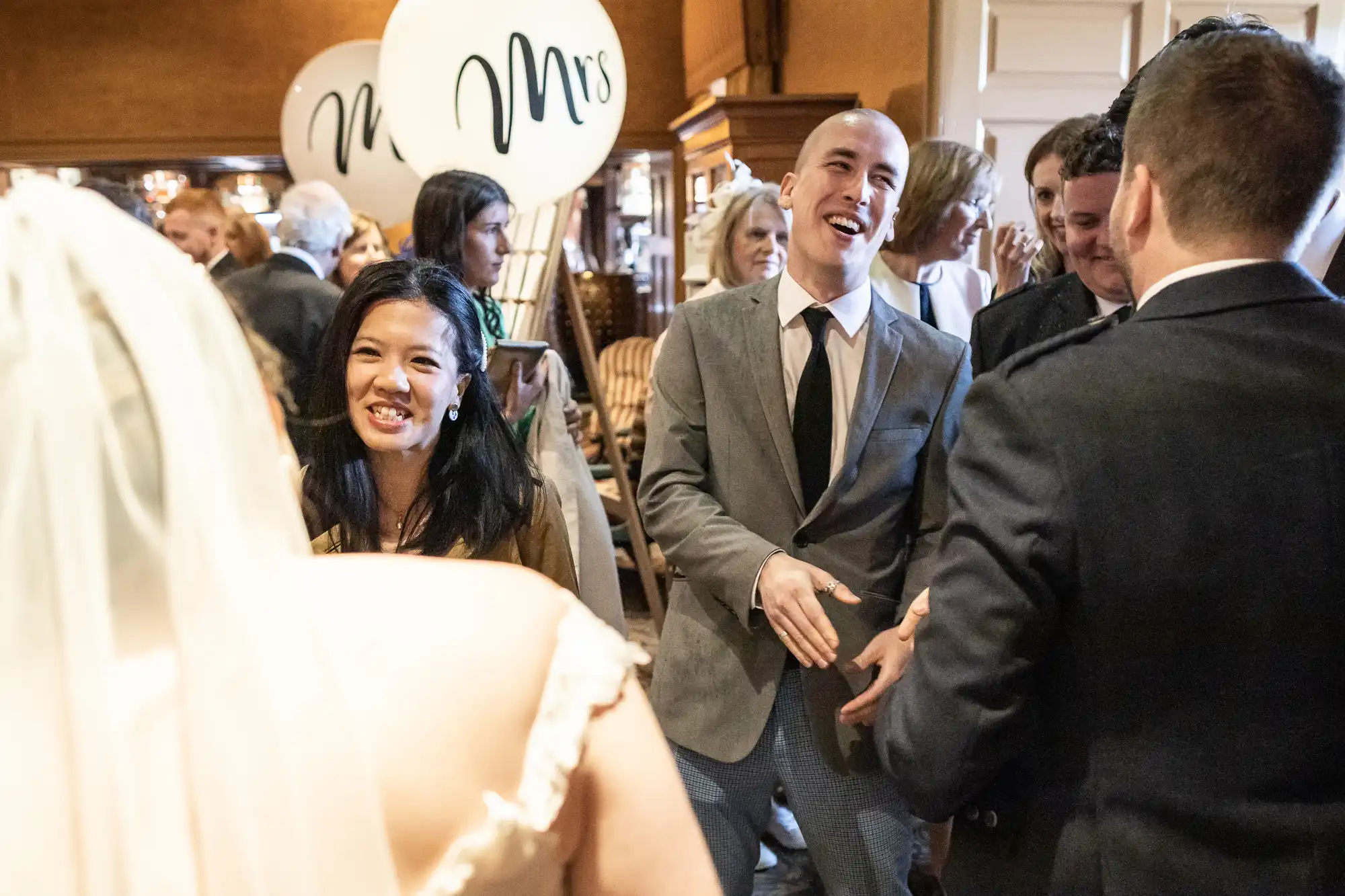 Guests at a wedding reception greet each other cheerfully; a bride with her back to the camera is in a white dress; one man extends a handshake. Large "Mrs" balloons are visible in the background.