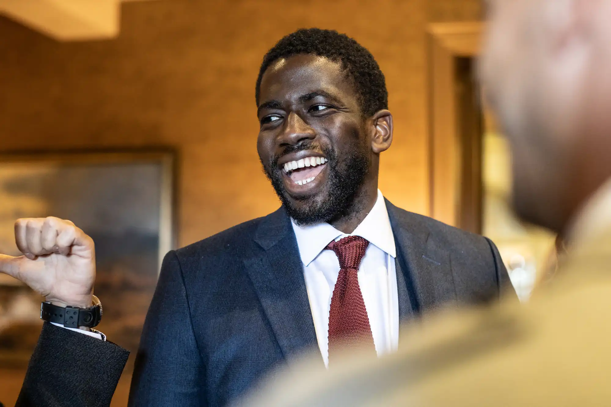 Man in a suit and red tie smiles while engaging in conversation with others at an indoor event.