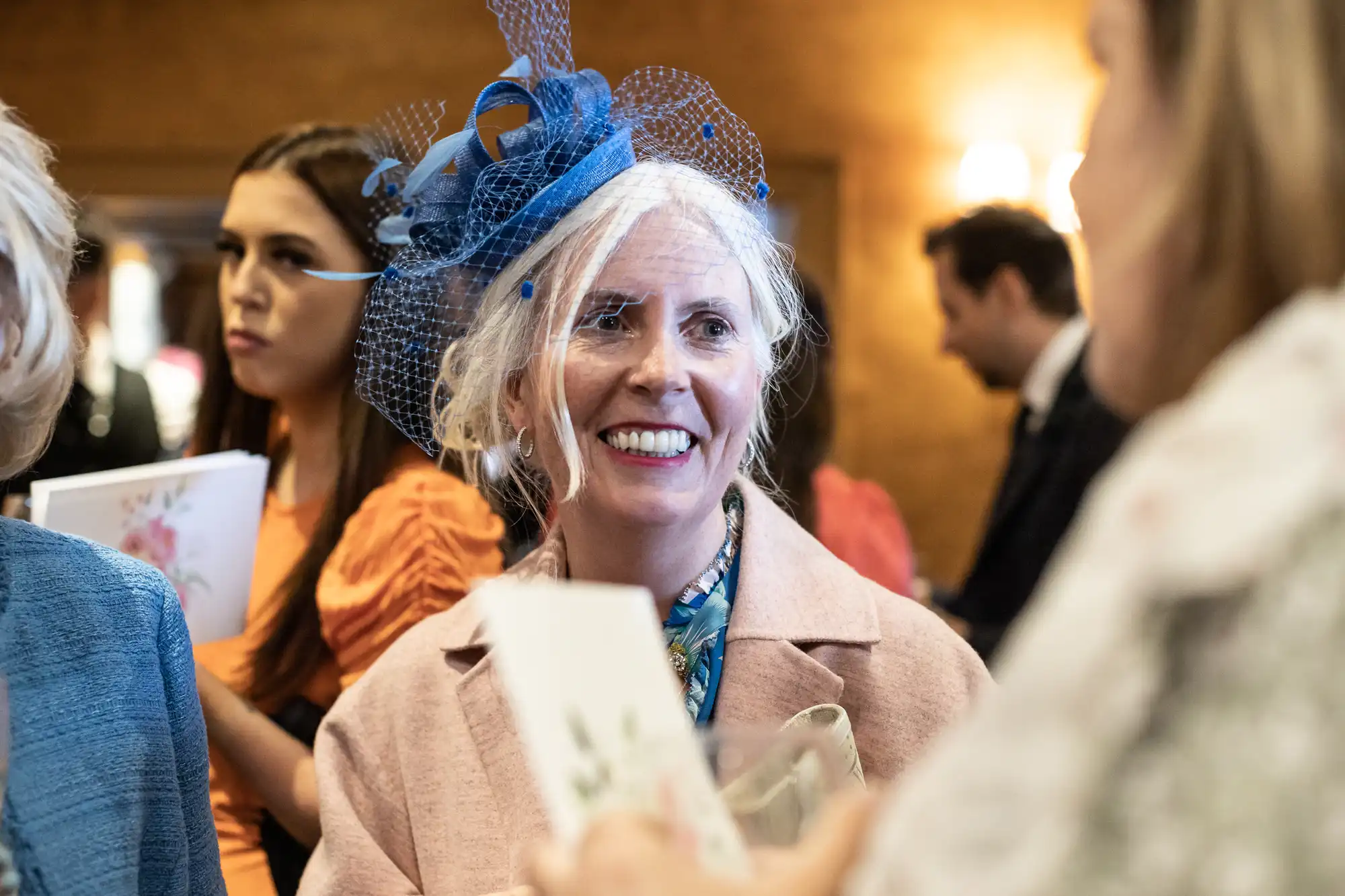 A woman with white hair wearing a blue fascinator smiles while holding a card at a formal event. She is engaged in conversation with another woman. Blurred people are in the background.