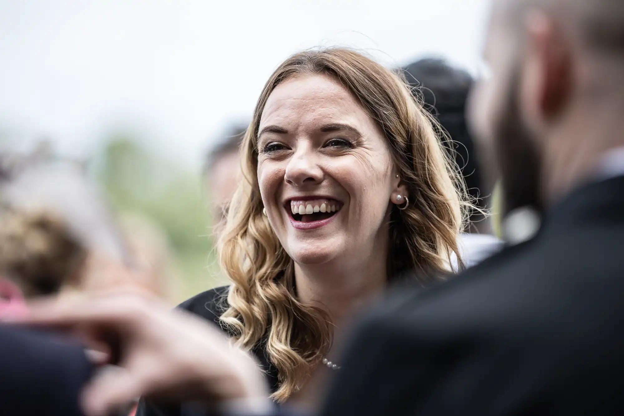 A woman with long blond hair, wearing a black top, is smiling and laughing in a crowd.