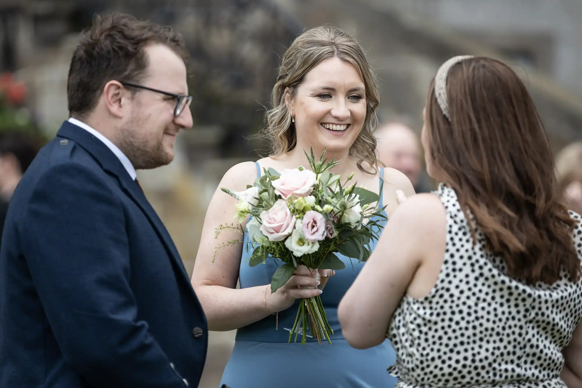 Three people are conversing outdoors. The woman in the middle is holding a bouquet of flowers and smiling at the woman on the right. The man on the left is also smiling.