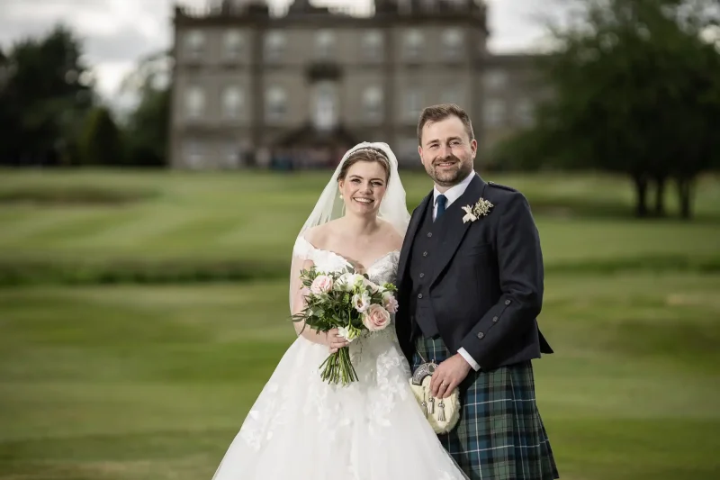 Wedding reception at the Dalmahoy Hotel: A bride in a white dress and a groom in a kilt pose together outdoors with a large building in the background.