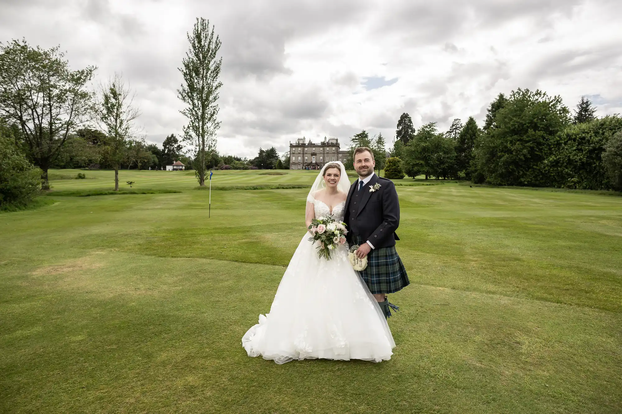 A bride in a white dress and a groom in a kilt and jacket pose together on a green lawn with trees and a large building in the background.